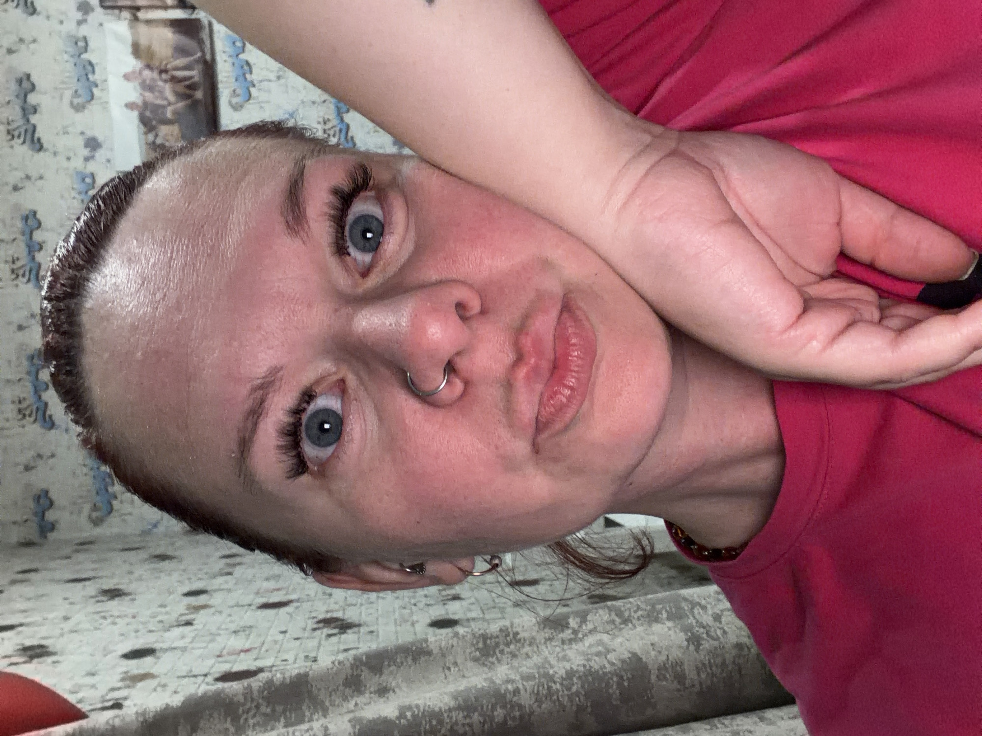 A close-up, rotated image shows a person with fair skin, light blue eyes, and dark eyebrows and eyelashes. They have a nose ring and multiple earrings in their left ear. The person is wearing a reddish-pink shirt. Their right arm is raised and bent, with their hand near their head. The background includes a patterned surface and what appears to be a curtain or wall hanging. There is no indication of the city of Chaikovsky, Russia in the image.