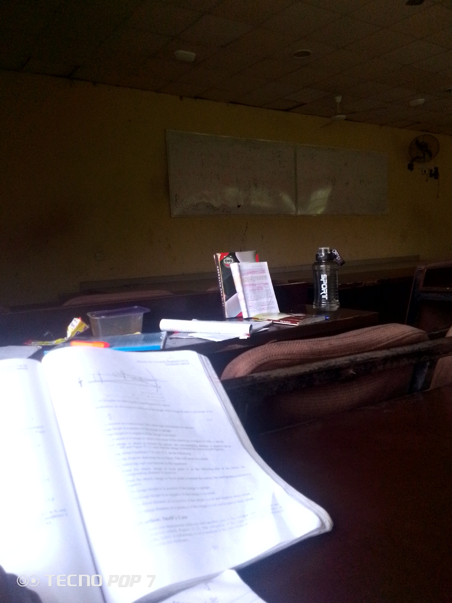 The image depicts an empty classroom setting. Two whiteboards are visible on a wall, one of which has some faint writing. In the foreground, a book lies open on a wooden desk, with some papers and a clear plastic container next to it. A black water bottle with "SPORT" written on it in white lettering stands on a desk in the middle ground. A ceiling fan is visible overhead, and a wall-mounted fan is also present on the right side of the room. The lighting suggests it may be daytime, with natural light filtering into the room. The overall impression is of a classroom that has been recently used or is prepared for a lesson, but is currently unoccupied. The image context places this scene in Eleweran, Nigeria.