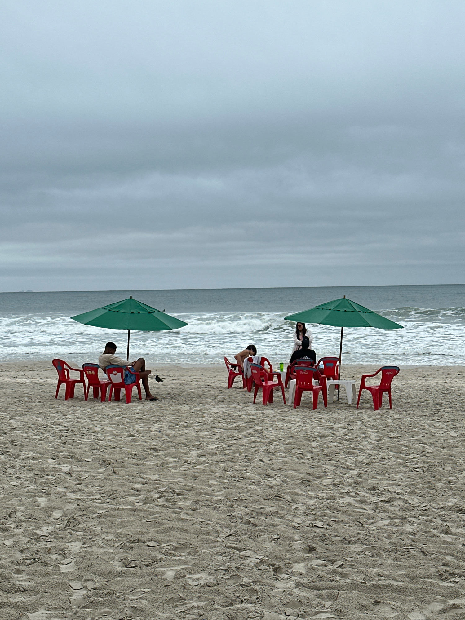 The image depicts a cloudy day at Praia dos Ingleses in Florianópolis, Brazil. Two groups of people are seated at tables under green umbrellas on a sandy beach. The group on the left consists of one person sitting in a red chair, facing away from the camera. The group on the right has at least three people visible, with one standing and two seated at a table with red chairs and a white table. The ocean with moderate waves is visible in the background under a heavily overcast sky. Several red plastic chairs and small tables are scattered on the sand. A bird is visible on the sand in the foreground of the left group.