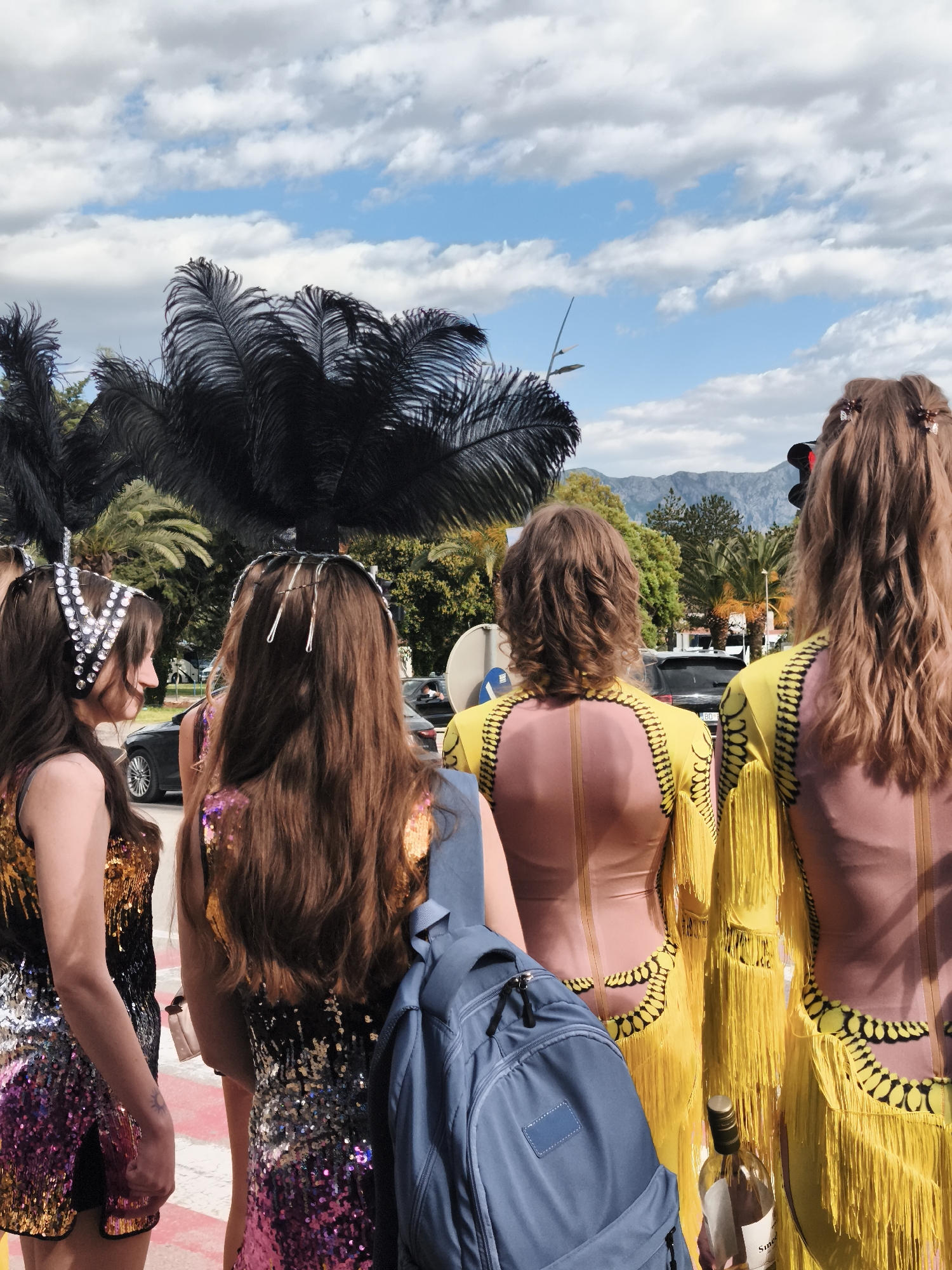 Four women stand on a red-and-white painted crosswalk at an outdoor location. The woman closest to the left wears a sleeveless dress covered in sequins, with a black headband decorated with white rhinestones. The woman next to her wears a similar dress in shades of pink and gold. She is wearing silver barrettes and carrying a blue backpack. Two women stand to the right, both wearing yellow dresses with long fringe. The dresses have pink panels on the backs and ornate black trim. The woman furthest to the right has hair adorned with small butterfly clips, and holds a bottle of wine. In the background there are trees, including black palm trees with feathery fronds, vehicles, and a mountain range in the distance under a blue sky with white, puffy clouds. A traffic light is visible.