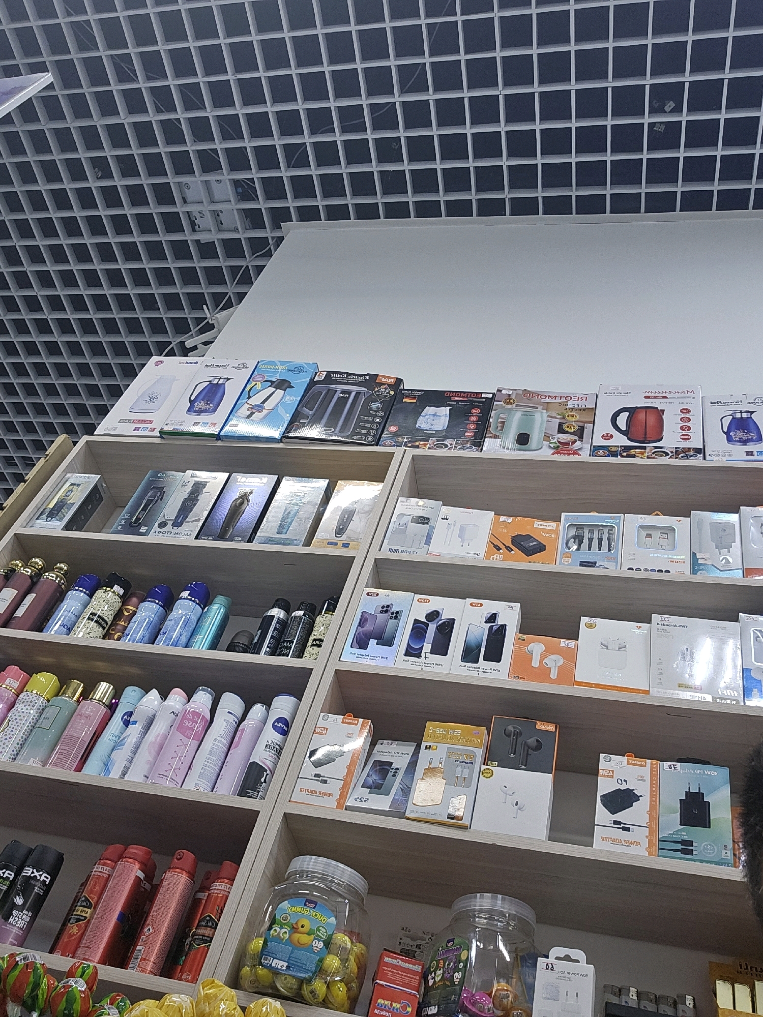The image depicts a retail store shelf filled with various consumer goods in Dushanbe, Tajikistan. The shelves are organized into sections, displaying items such as electric kettles, hair clippers, personal care products like deodorants and perfumes, phone accessories such as chargers and headphones, and packaged candies. The store appears well-lit, with a white grid ceiling visible in the upper portion of the image, typical of commercial spaces. The products are arranged in their original packaging, indicating a well-stocked retail environment.
