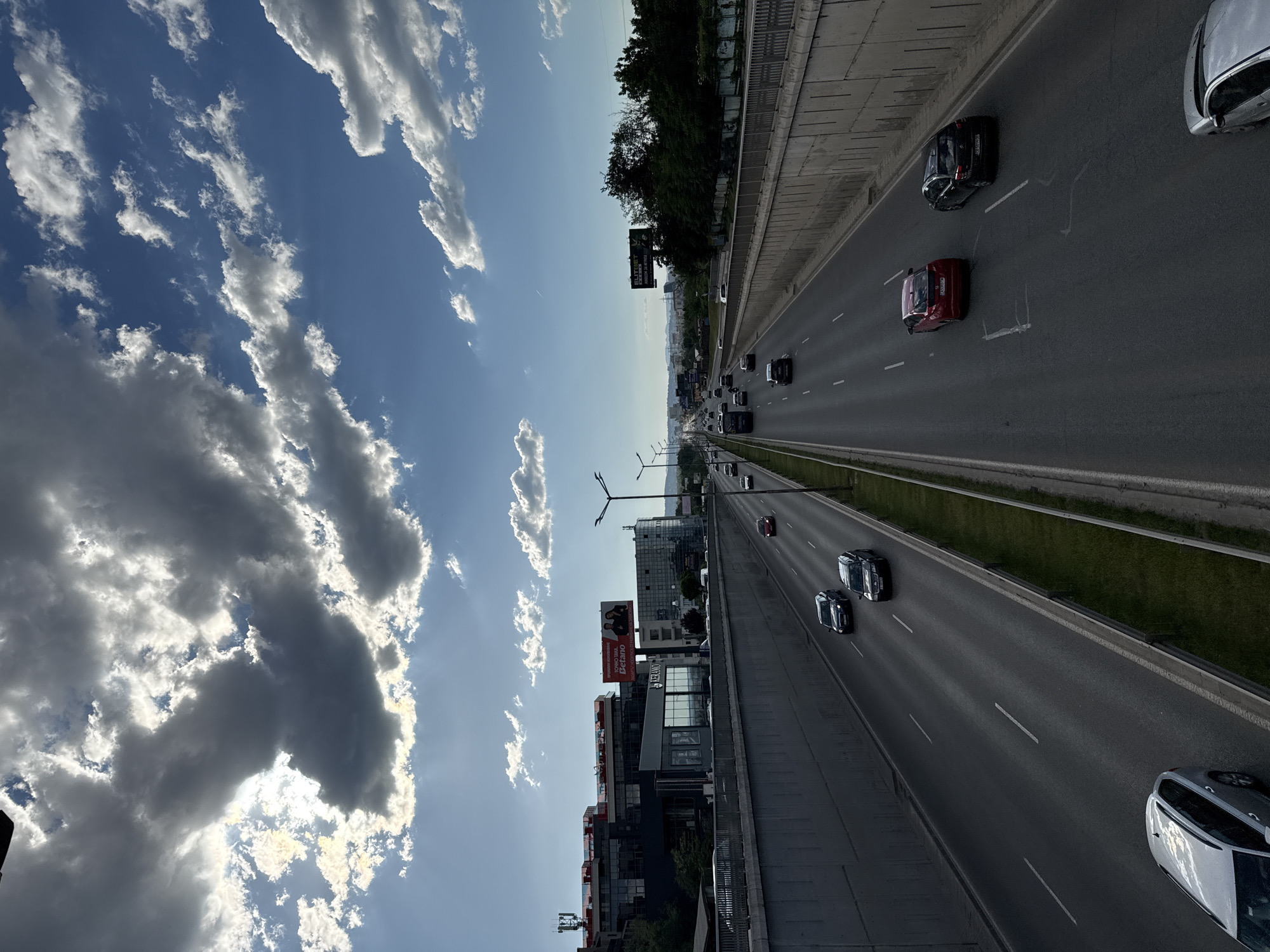 The image is a high-angle, daytime shot of a highway in Sofia, Bulgaria. The highway, featuring multiple lanes of traffic, stretches into the distance, with cars visible in both directions. On one side of the highway is a cityscape with modern buildings and billboards. The sky is filled with cumulus clouds and a bright blue sky. The overall impression is of a busy urban environment on a sunny day.
