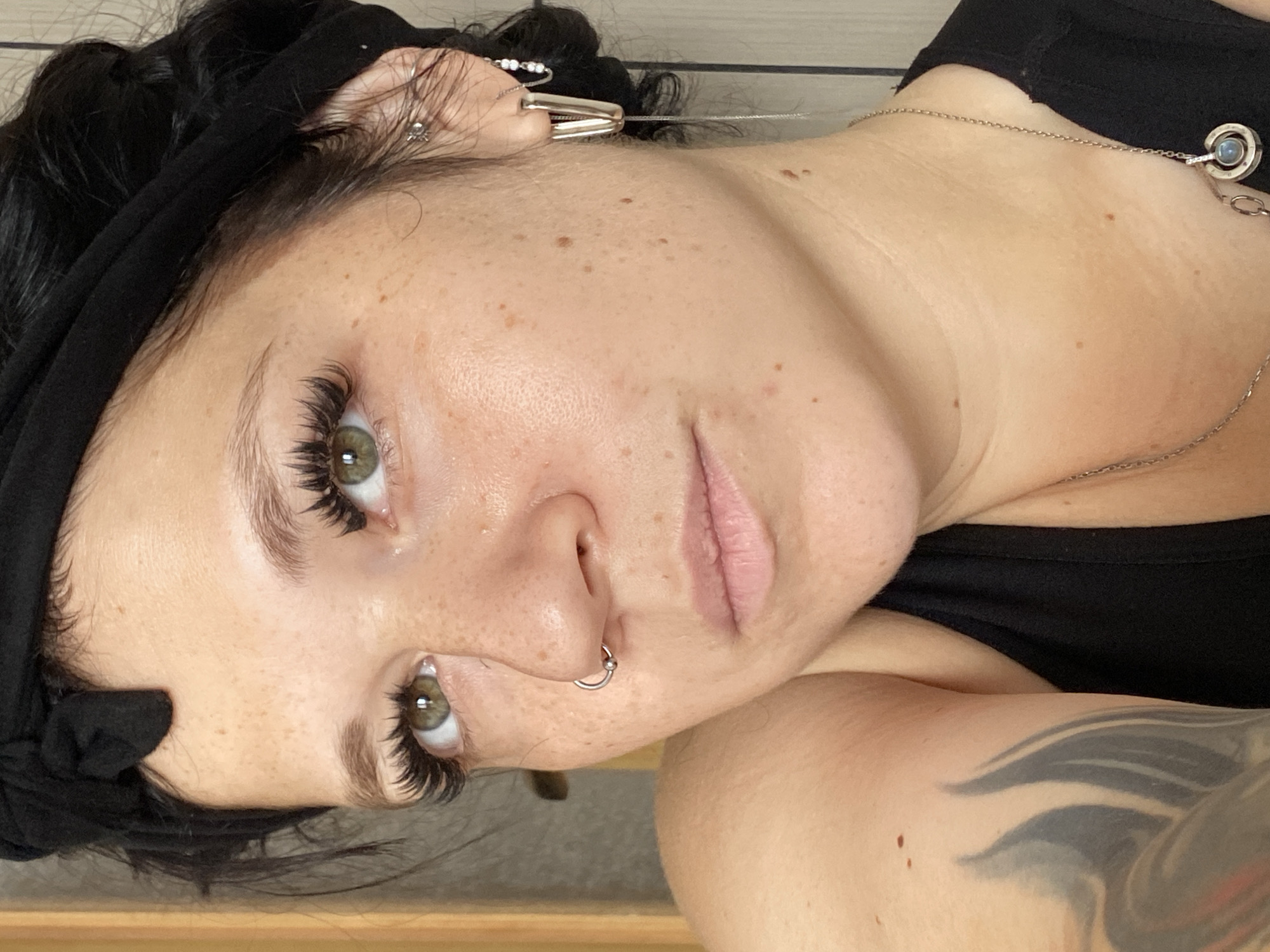 The image is a close-up portrait of a woman with freckles and green eyes, looking slightly downwards and to her left. She has on false eyelashes, a black headband tied at the top of her head, and a nose ring. She is wearing a black tank top, and a silver necklace with a pendant. Her left arm, partially visible, has a large tattoo. The background is out of focus and appears to be an indoor setting with light-colored vertical blinds on the right. The lighting suggests it is daytime. The image is cropped to focus on the woman's face and upper body. There is no information in the image to suggest its location in the city of Ostrovki, Russia.