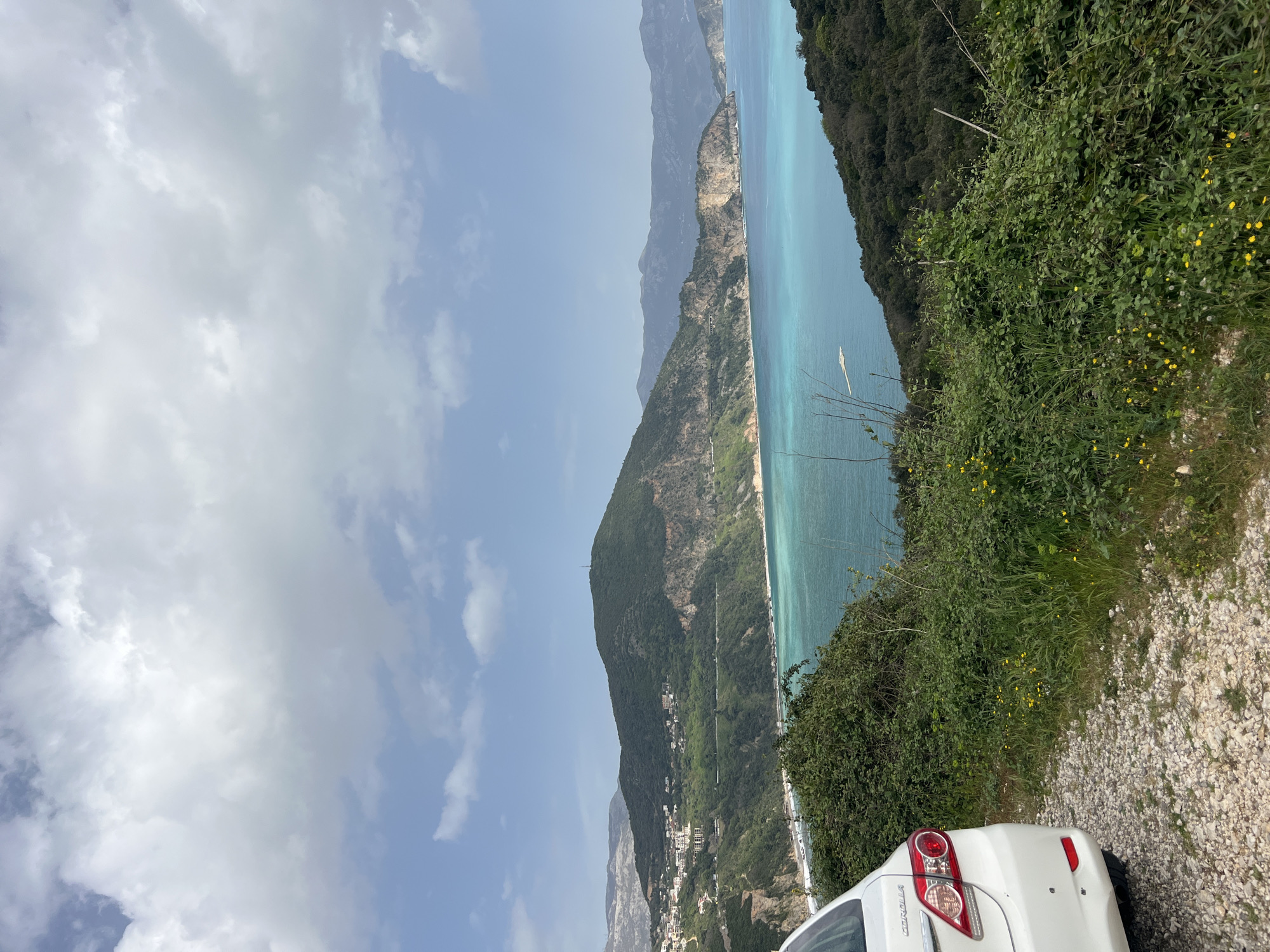 The image depicts a coastal view in Visnjeva, Montenegro, taken from an elevated perspective next to a road. To the right, a white car, possibly a Toyota Corolla, is partially visible, parked on a gravel shoulder. Lush green vegetation, including trees, bushes, and yellow wildflowers, covers the slope. The coastline stretches into the distance, with mountains rising along the coast. A narrow beach runs along the base of the mountains. The sea is a clear turquoise color, and there are some boats visible on the water. The sky is light blue with scattered white clouds. The image seems to have been rotated, as the sky is on the left.