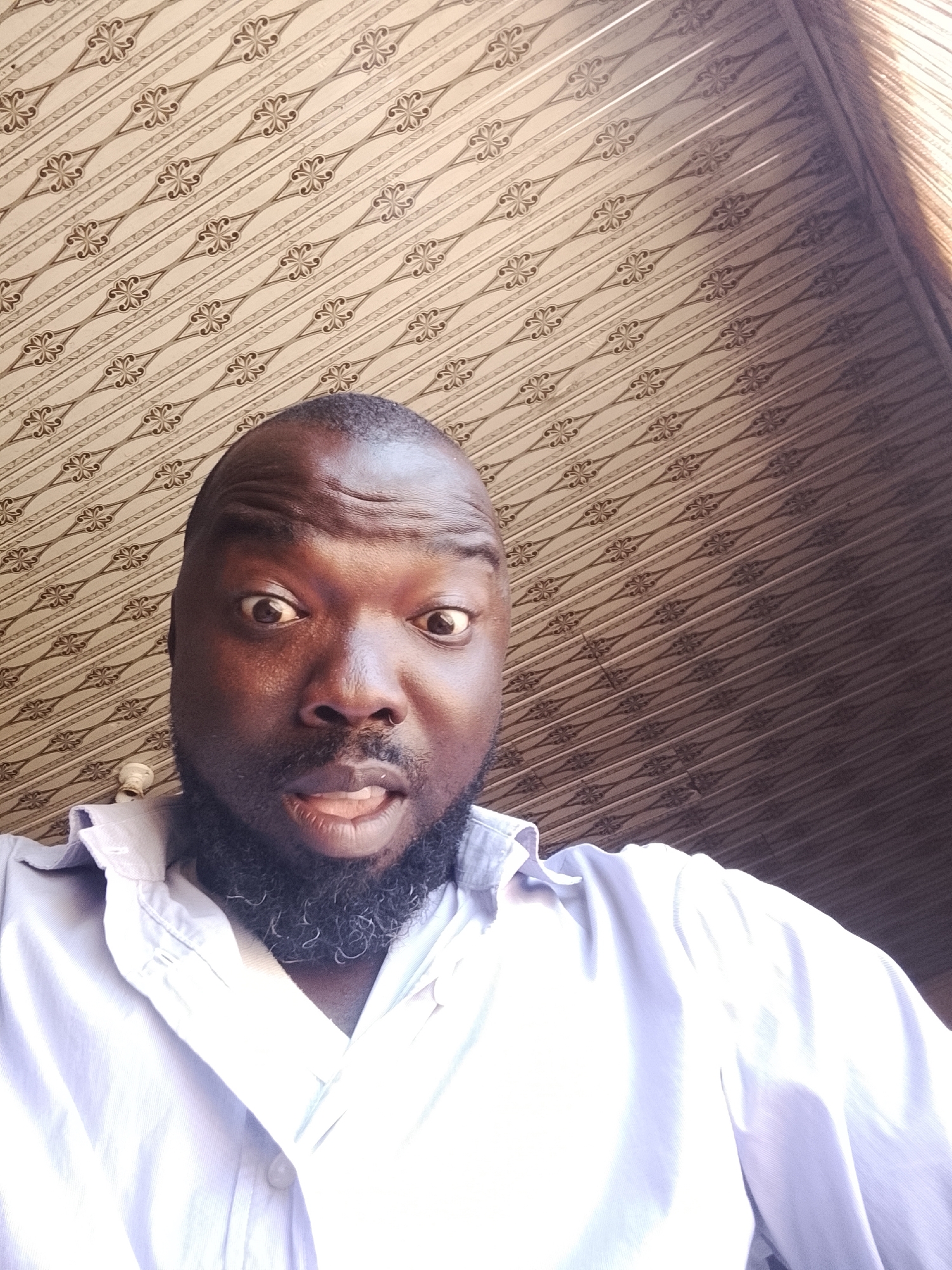 The image shows a medium close-up of a Black man with a shaved head and a full black beard, wearing a light blue button-down shirt. He is looking directly at the camera with a surprised expression, his eyes wide and mouth slightly open. The background features a patterned ceiling with a repetitive design of beige and brown floral elements arranged in diagonal rows. The location is Gwong, Nigeria, based on context outside of the image itself.