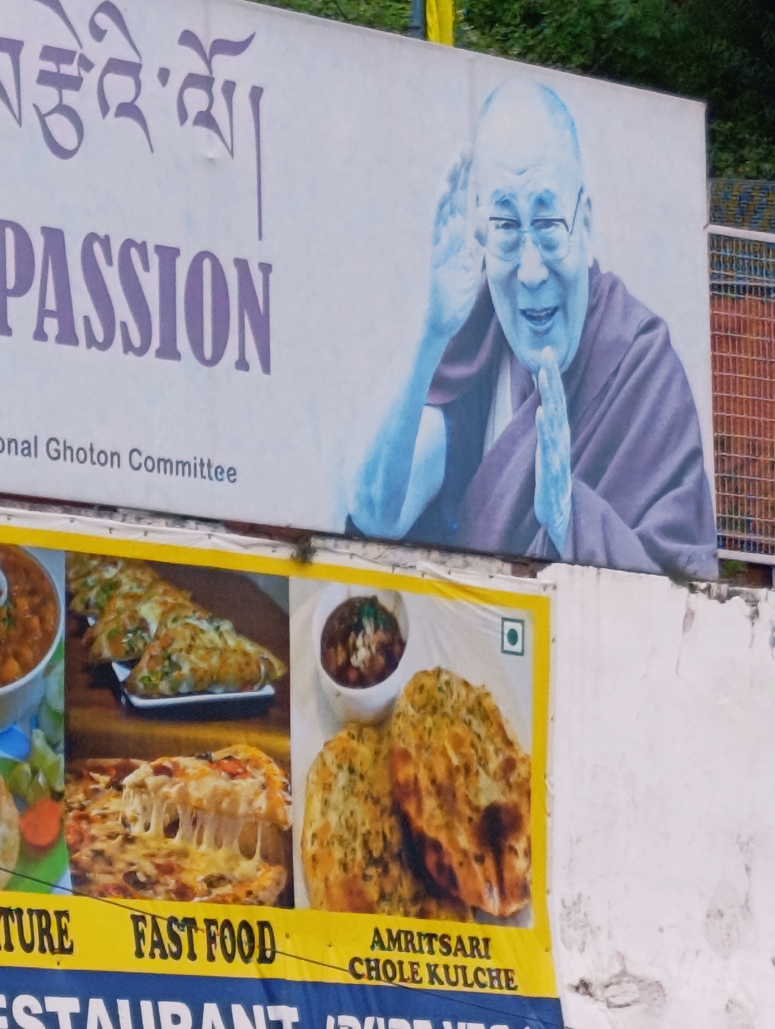 The image displays a large billboard in Kalath, India, featuring a portrait of the Dalai Lama on the right side. The billboard also has Tibetan script at the top and the word "PASSION" in large, purple letters. Below "PASSION," the text "onal Ghoton Committee" is visible. The lower portion of the billboard contains images of food, including pizza, what appears to be naan or kulcha, and bowls of curry or dal. A yellow banner runs along the bottom, with text that includes "URE FAST FOOD" and "AMRITSARI CHOLE KULCHE". The billboard is mounted on a white wall, and some of the food images are bordered in yellow. The background shows greenery and a brick structure on the right.