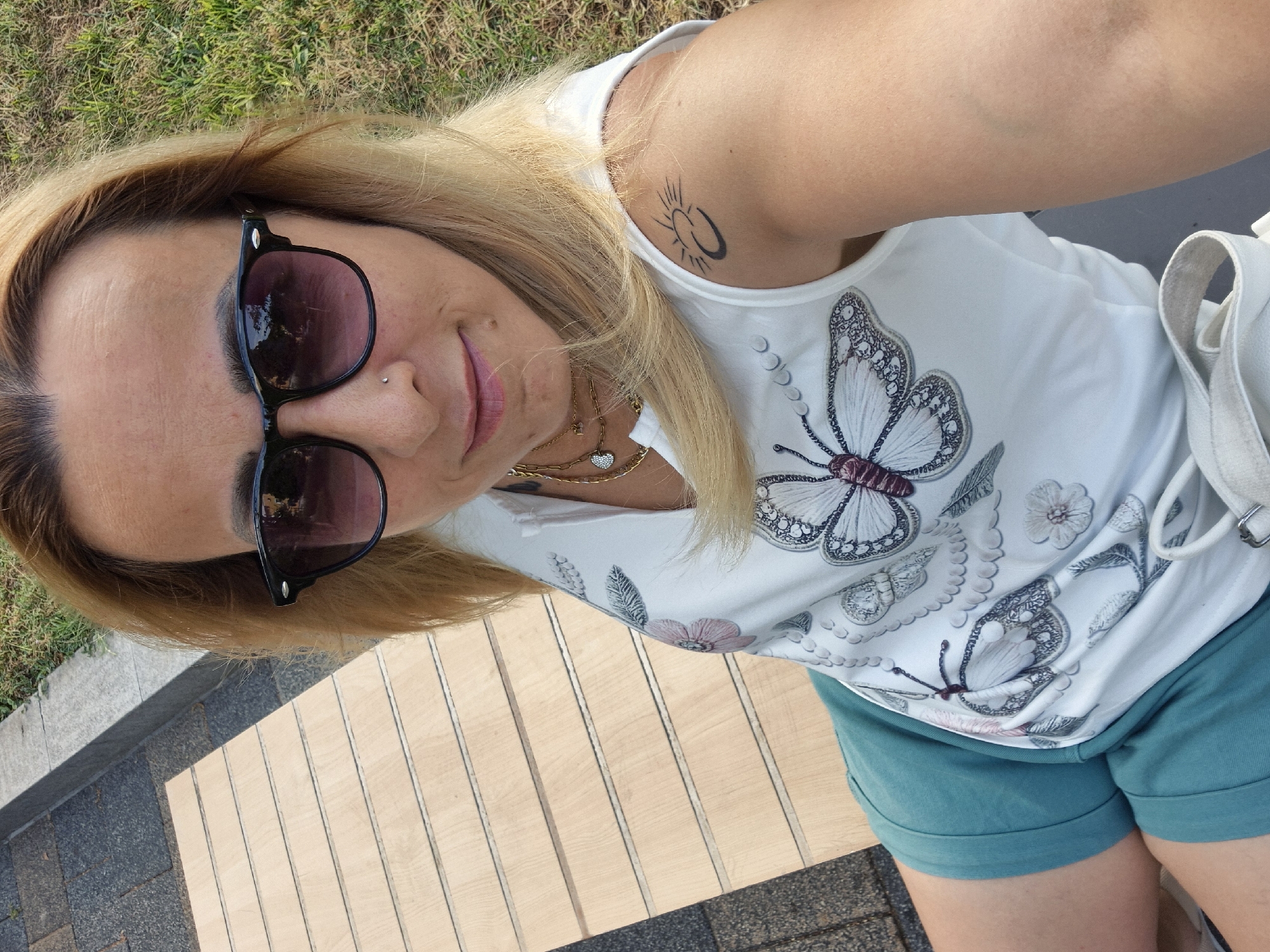 An outdoor, high-angle, medium selfie shows a woman with blonde hair, wearing sunglasses and a white tank top with a butterfly print, sitting on a wooden bench in an urban park. She is wearing teal shorts. A tattoo of a crescent moon and sun is visible on her left shoulder. The bench is situated next to a paved walkway and a grassy area.