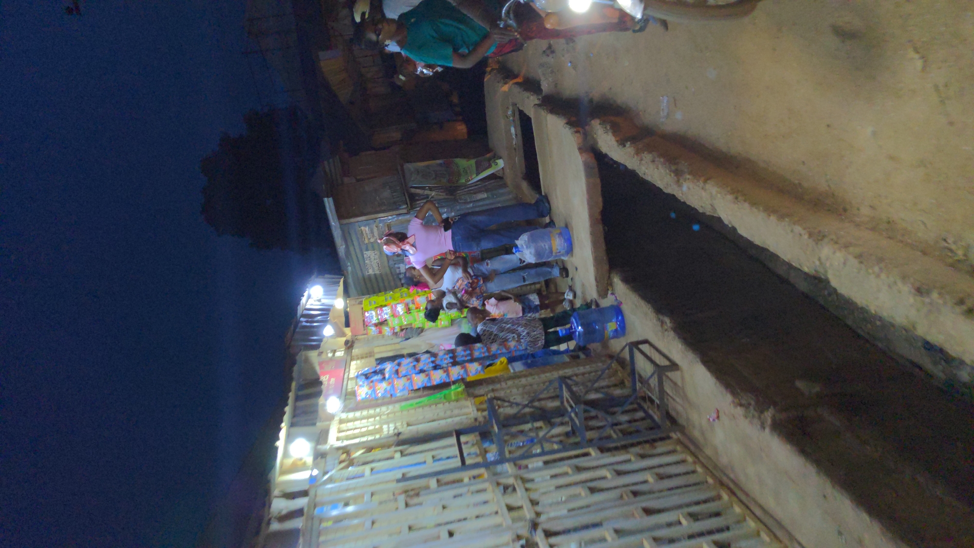At dusk in Ado, Nigeria, a street vendor's stand is set up beside a concrete drainage ditch. The stand, illuminated by several bright lights, displays snacks and other goods. A group of people stand near the stand, some holding large blue water jugs, suggesting they are purchasing water. Several other people are present on the other side of the road.