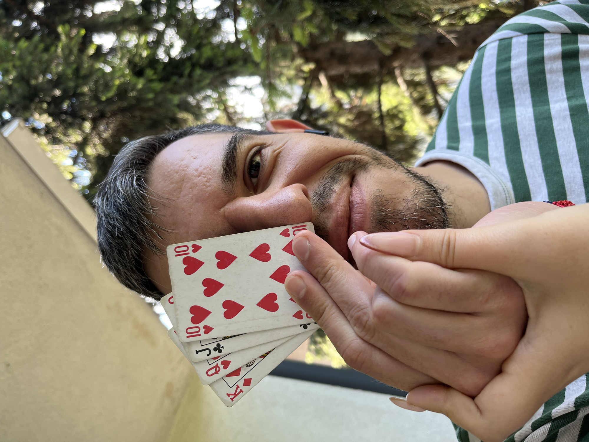 A man with graying hair and a goatee holds four playing cards to his right eye, obscuring it. The cards are the King of Diamonds, Queen of Hearts, Jack of Clubs, and two 10 of Hearts. His left hand is clasped over his right, with a person's fingers and thumb visible, supporting the cards. The man wears a green and white striped t-shirt. He is outdoors, with a blurred background of green foliage, suggesting trees. The image appears to be taken on a sunny day. The scene is a casual outdoor setting. There is no identifiable location in Sofia, Bulgaria.
