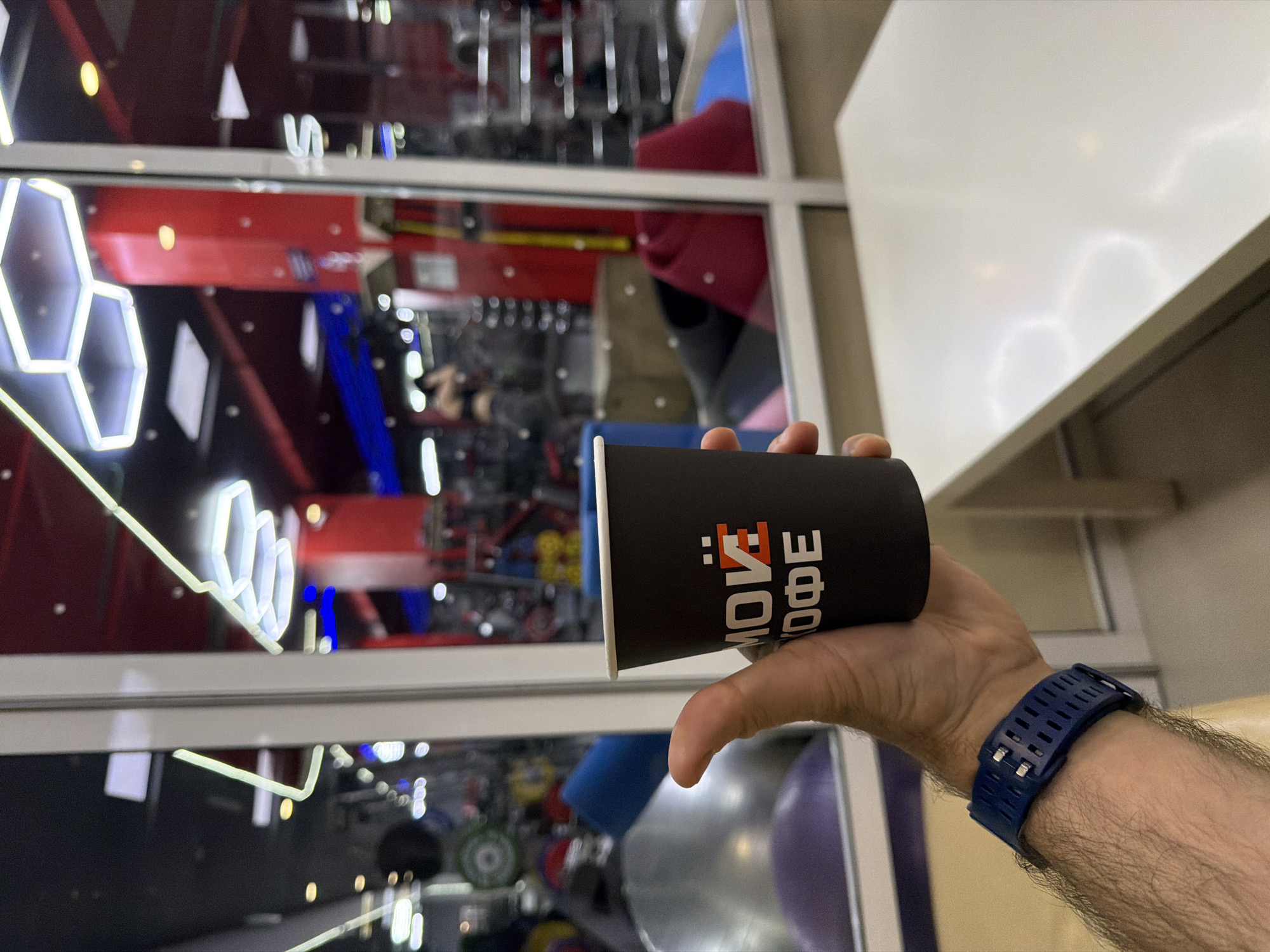 A first-person perspective shot shows a person's arm and hand holding a black paper cup labeled "Moi Coffee" in Cyrillic. The person wears a blue sports watch. The background shows a bright, modern gym or sports facility interior through a window. The facility has climbing walls with various colored holds, illuminated geometric designs, and fitness equipment. The location is likely Krasnoyarsk, Russia, based on the cup's branding.