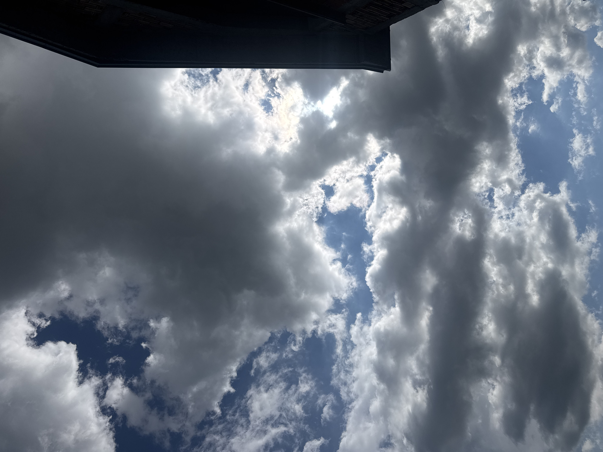 Looking upwards, the sky is filled with a mix of dark, ominous storm clouds and patches of bright blue sky. The clouds are various shades of gray and white, with the darker formations hinting at an impending storm. Sunlight is partially obscured by the dense cloud cover. In the upper portion of the frame, part of a dark building structure is visible, partially obscuring the sky. No people or objects are visible. The photo does not reveal if the image was taken in Sarnevo, Bulgaria.