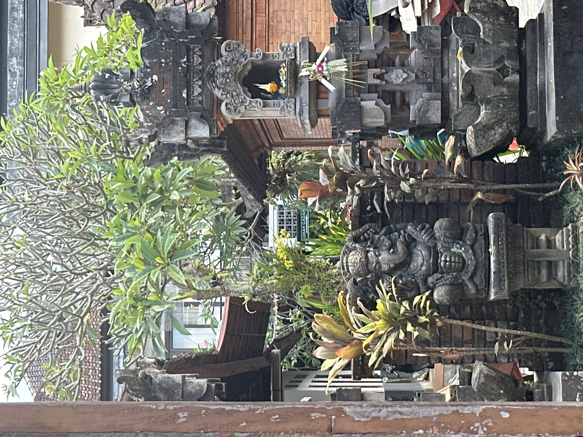 An outdoor scene in Ubud District, Indonesia, features a stone statue of Ganesh, the Hindu deity with an elephantine head, positioned prominently in the foreground. The statue is seated on a carved pedestal and is surrounded by lush green foliage and a brick wall. To the right of the Ganesh statue, a smaller stone shrine is visible, adorned with offerings. Beyond these elements, traditional Balinese architecture with tiled roofs and intricate stonework is evident. The overall atmosphere suggests a cultural or religious setting within a tropical environment.