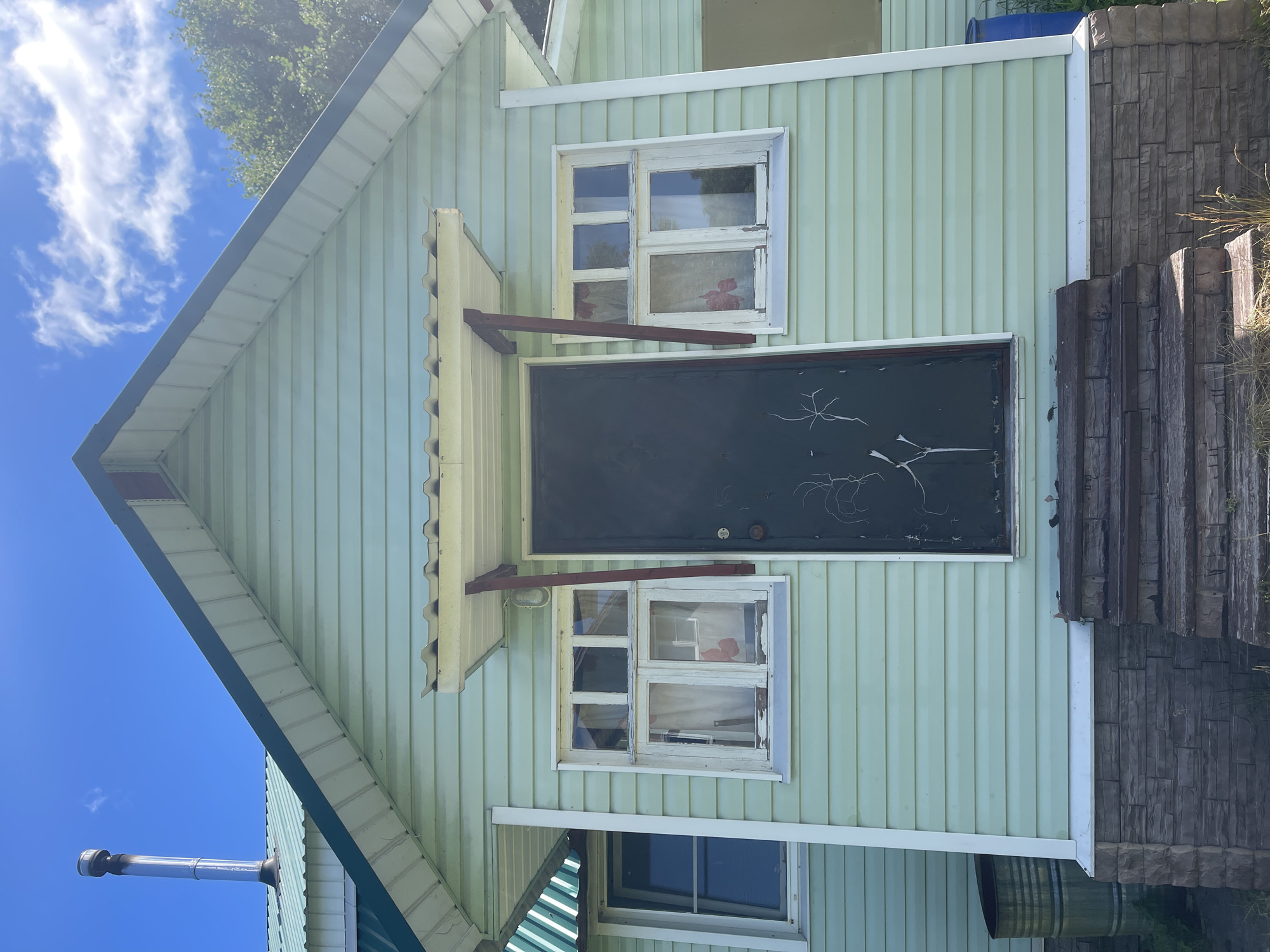 This is a front-facing, eye-level, medium shot of the light green, horizontal vinyl-sided facade of a small, single-story house under a bright blue sky with scattered white clouds. The house has a light green metal roof with a silver metal chimney pipe visible on the left side of the roof. There is a dark door in the center of the house, flanked by two white-framed, four-pane windows on each side. Both windows have white curtains visible. Above the door is a small, white, corrugated metal awning supported by two dark red, angled beams. The foundation of the house is covered with gray stone veneer. Wooden steps, showing signs of wear, lead up to the door. To the left of the steps, a large, green metal barrel is visible, and to the right, a blue plastic barrel is partially visible. The house appears to be located in a rural or suburban area, as indicated by the overgrown grass and weeds in front of the house and the trees visible in the background on the right side. The overall condition of the house suggests it is older and may require some maintenance. The location is identified as Krasnoye, Russia.