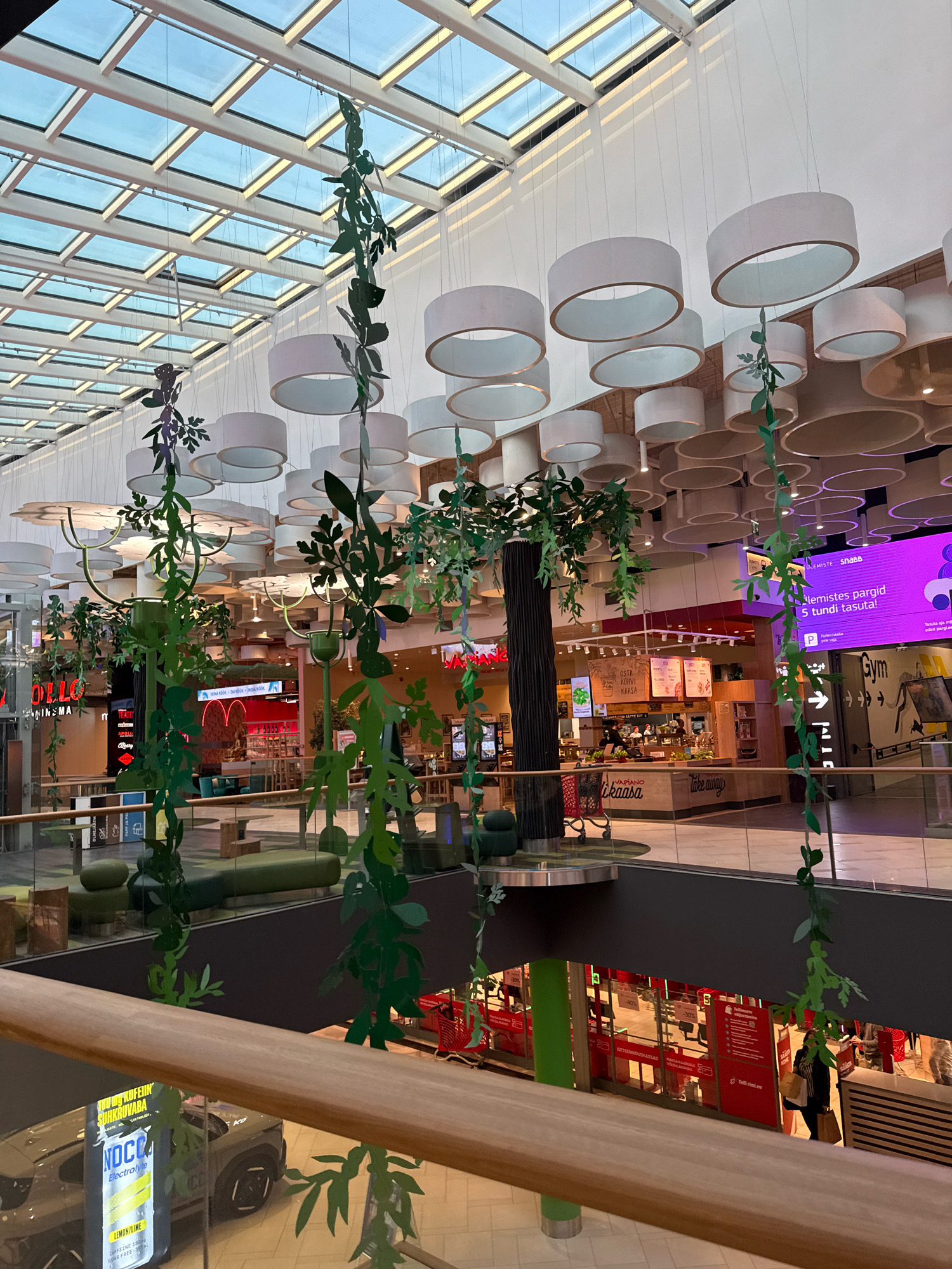 The image shows the interior of the Ülemiste Centre, a shopping mall in Tallinn, Estonia. The perspective is from an upper level, looking down towards the ground floor and across to various shops and eateries. A prominent feature is the ceiling, which has a glass roof with a grid of white metal supports and numerous large, white cylindrical pendant lights.
Green decorative foliage hangs from the ceiling and from artificial trees, adding a natural element to the modern mall architecture. On the ground floor, several shops and food vendors are visible. One food stall has "OSTA KÕRVALT" and "VÄRSKE KÄSITÖÖ SAIAD" signs. Another area has a "Take Away" sign and appears to be a restaurant or cafe. In the background, a large purple digital screen displays text, including "ÜLEMISTE SNABB" and "Telli tasuta". A sign for "Gym" is also visible.
In the foreground, a wooden railing separates the upper level from the lower levels. Below the railing, part of a car and a display for a product labeled "NOCO Electrolyte" are visible. People are present on the ground floor, including a woman carrying shopping bags walking past a store with red signage and discount percentages displayed. The overall environment suggests a busy, well-lit, and modern shopping complex.