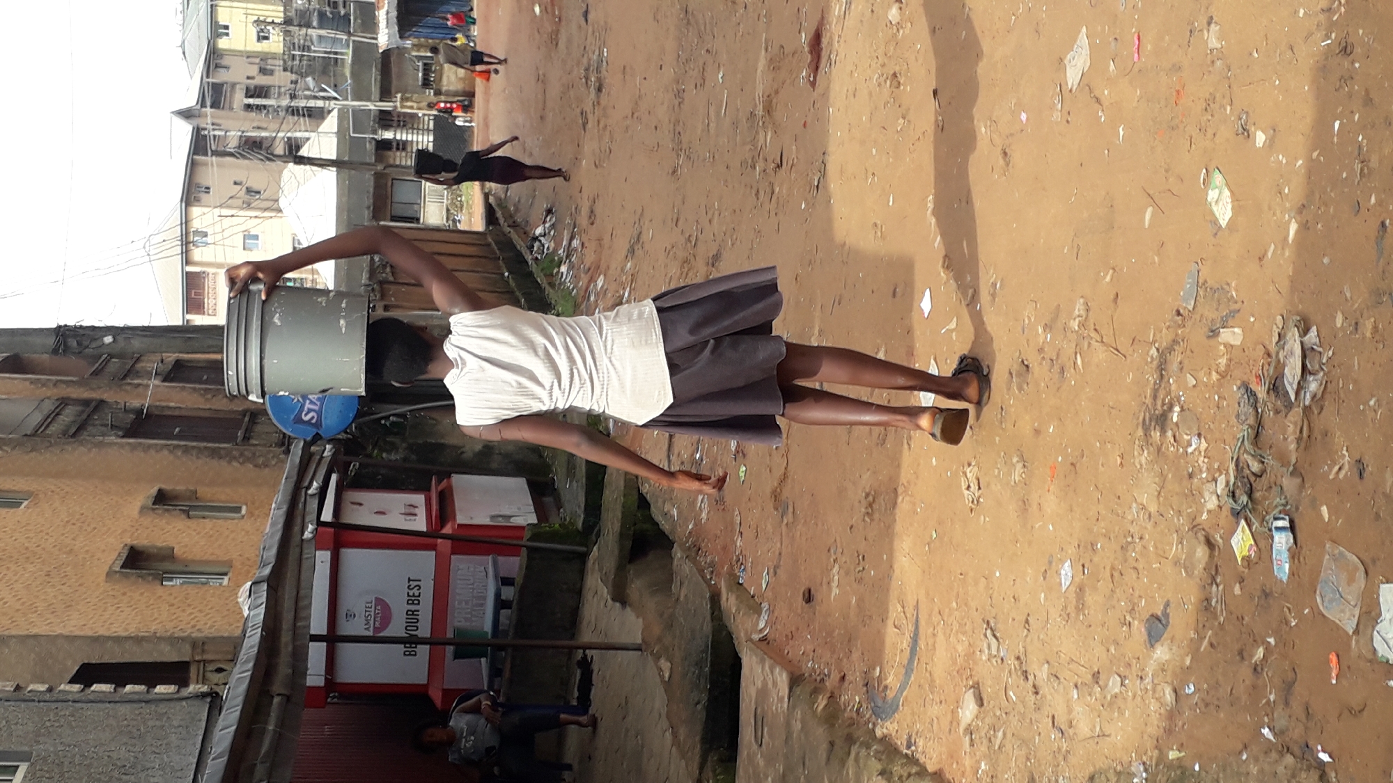 A young woman, viewed from the rear, walks down a dirt road in Nkpor, Nigeria, carrying a large gray bucket of water on her head. She is wearing a sleeveless white top and a dark, knee-length skirt. Her arms are bent at the elbows, with her right arm raised to balance the bucket. In the background, other individuals are walking, and buildings line the sides of the road, some appearing to be shops or residences. The ground is uneven and appears to be littered with debris. A blue sign with white lettering, possibly advertising "STAR," is visible on the left.