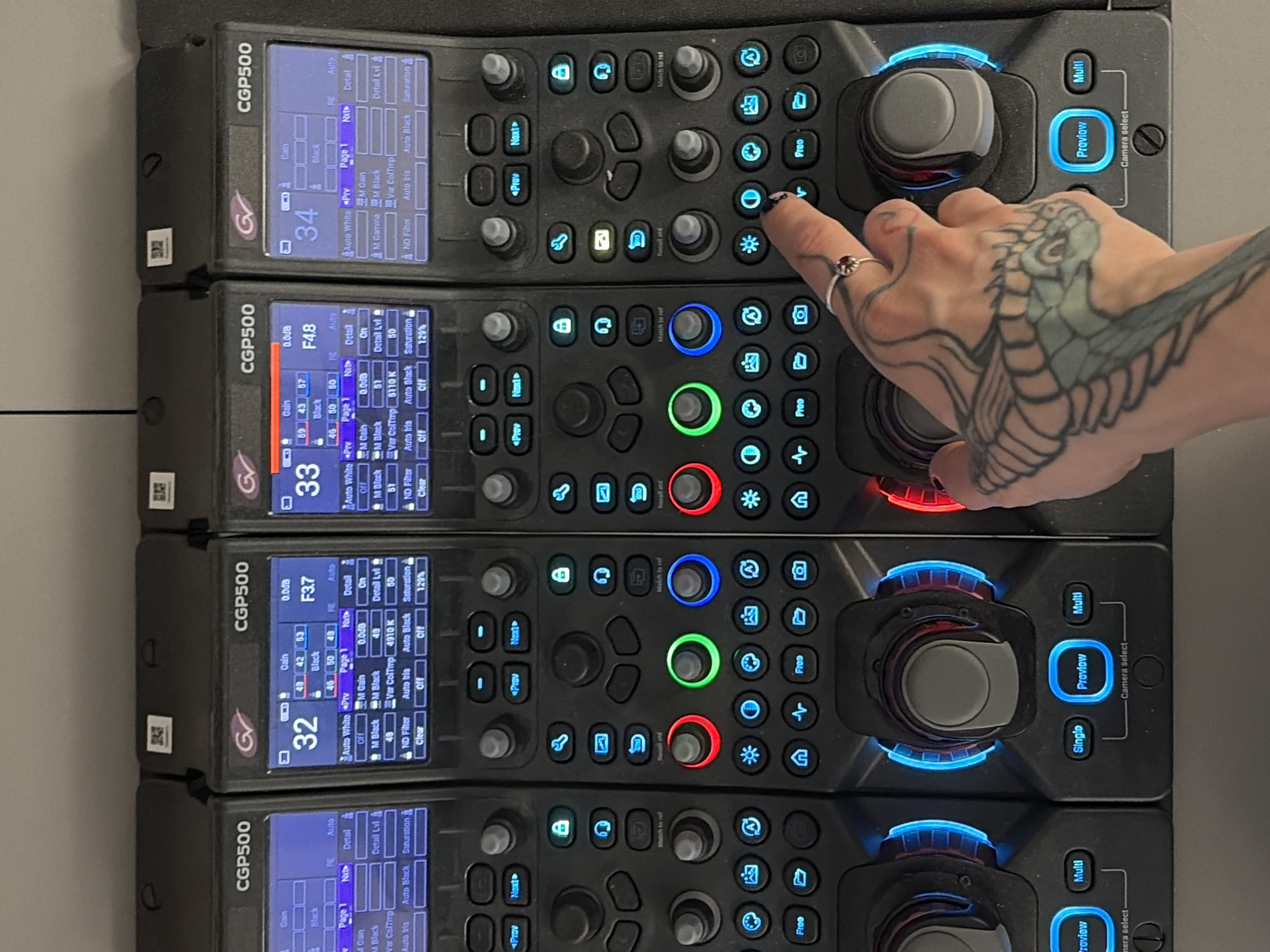The image shows a close-up view of four vertically stacked Grass Valley CGP500 camera control panels. Each panel has a screen displaying camera settings and a series of buttons and knobs for camera adjustments. A person's hand, with a snake tattoo, is pressing one of the illuminated buttons on the top panel. The control panels are black with blue, green, and red illuminated buttons. The location is unspecified and there is nothing in the image to suggest Sofia, Bulgaria.