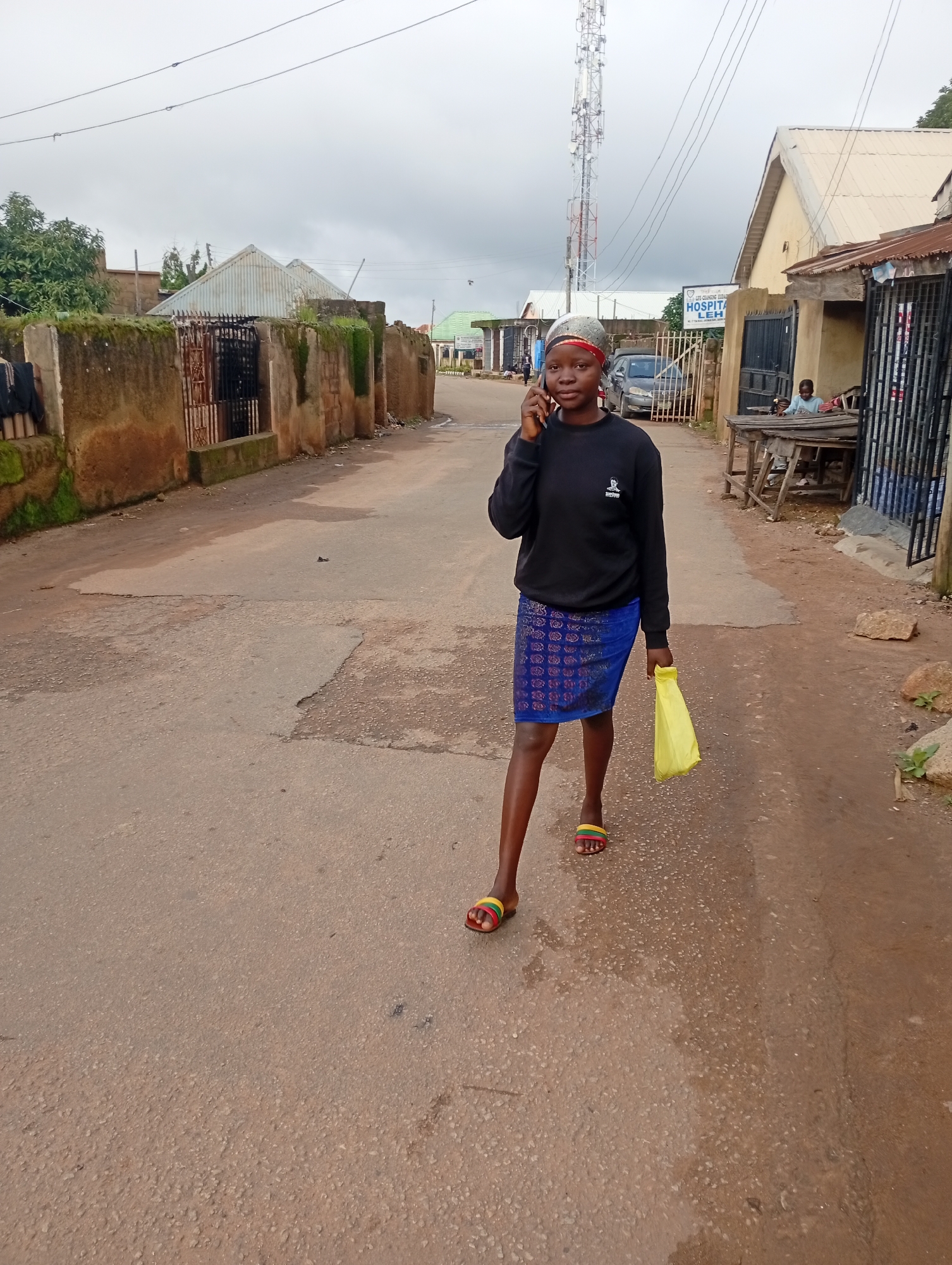 In Jos, Nigeria, a young woman, wearing a black sweatshirt and a blue patterned skirt, stands on a paved road while talking on a cell phone. She is carrying a bright yellow plastic bag in her right hand. Her feet are adorned with colorful slide sandals. Behind her, a cell tower dominates the skyline under a cloudy sky. To her right, a building with a sign reading "HOSPITAL LEH" is visible. Further down the road, a person sits at a wooden table outside a shop. The street is lined with various structures, including a stone wall with vegetation growing on it to the left, and more dilapidated buildings to the right. The road surface shows signs of wear and tear, with patches of broken asphalt and water stains. The overall atmosphere suggests an urban or semi-urban environment in Nigeria.