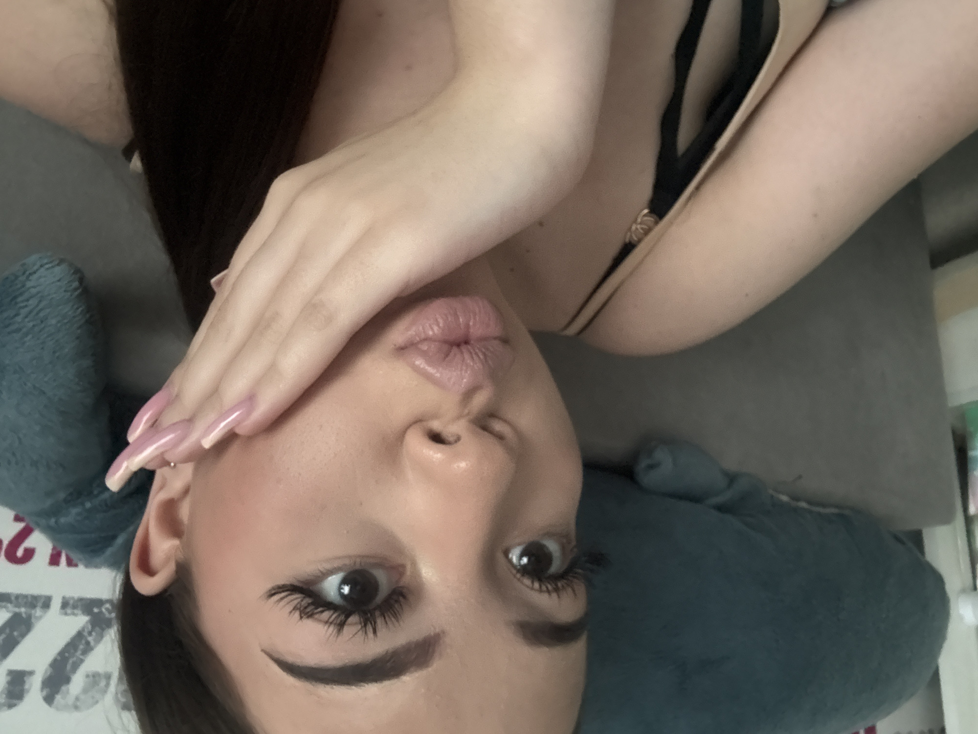 The image shows a woman's face, slightly angled, with her hand partially covering her forehead. She has dark hair, long eyelashes, and is wearing makeup. She is lying on a blue pillow or cushion, and part of her bra strap is visible. The background is blurry, but a calendar can be seen with the number 22 and 23 visible. There is insufficient information to determine the exact location in Naberezhnye Chelny, Russia.