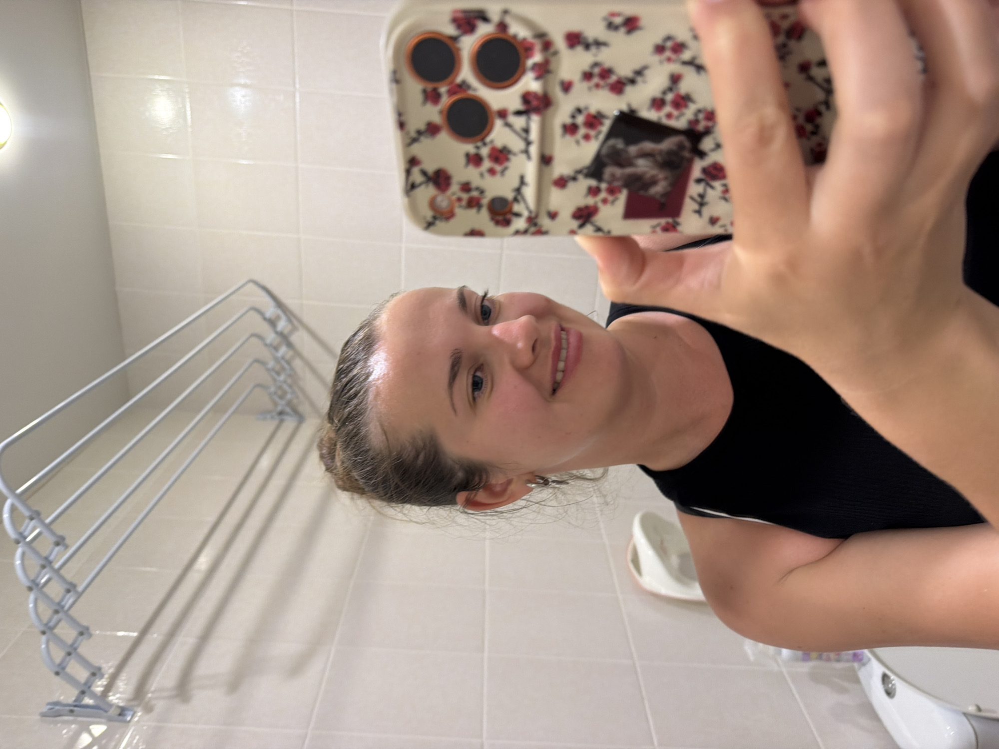 This media file is a photograph taken indoors, likely in a bathroom, as indicated by the tiled walls and floor, a drying rack, and a toilet. The image is rotated 90 degrees counter-clockwise.
The primary subject is a young woman who is holding a smartphone in her right hand, taking a selfie. She is smiling and looking directly at the camera. She is wearing a black tank top. Her hair is pulled back.
In the background, a metal drying rack with multiple bars is visible against the tiled wall. A toilet is partially visible in the lower right portion of the image. The room appears to be well-lit, suggesting it's daytime or the lights are on. There is no visible text in the image. The overall atmosphere appears casual and personal. The location context provided indicates the setting is Sochi, Russia.