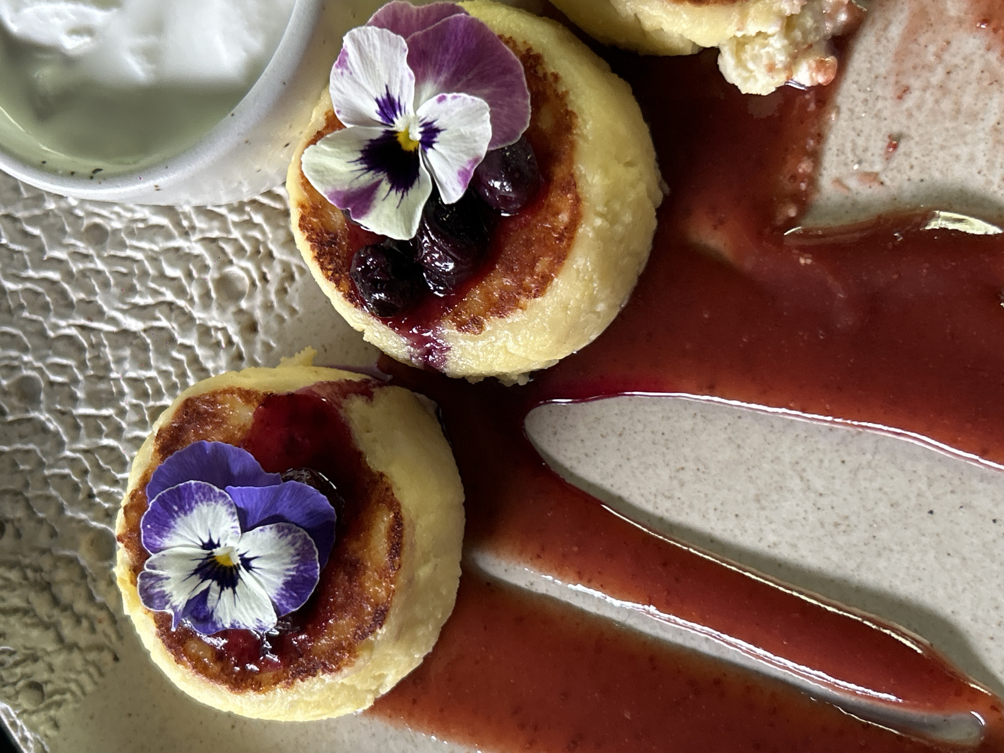 The image depicts a close-up of a plate of food. There are two small, round pancakes or fritters, each topped with a purple and white edible flower and what appear to be dark berries and possibly some kind of syrup or sauce. A separate bowl or container of white cream or yogurt is also on the plate. A dark red sauce is drizzled or swirled around the food on the plate. The plate has a light tan or beige base.
This is most likely food served at a restaurant, possibly in North Kuta, Indonesia. The specific type of food is not immediately evident, but the presentation suggests a carefully prepared dish, possibly a breakfast or dessert item. There are no people visible.