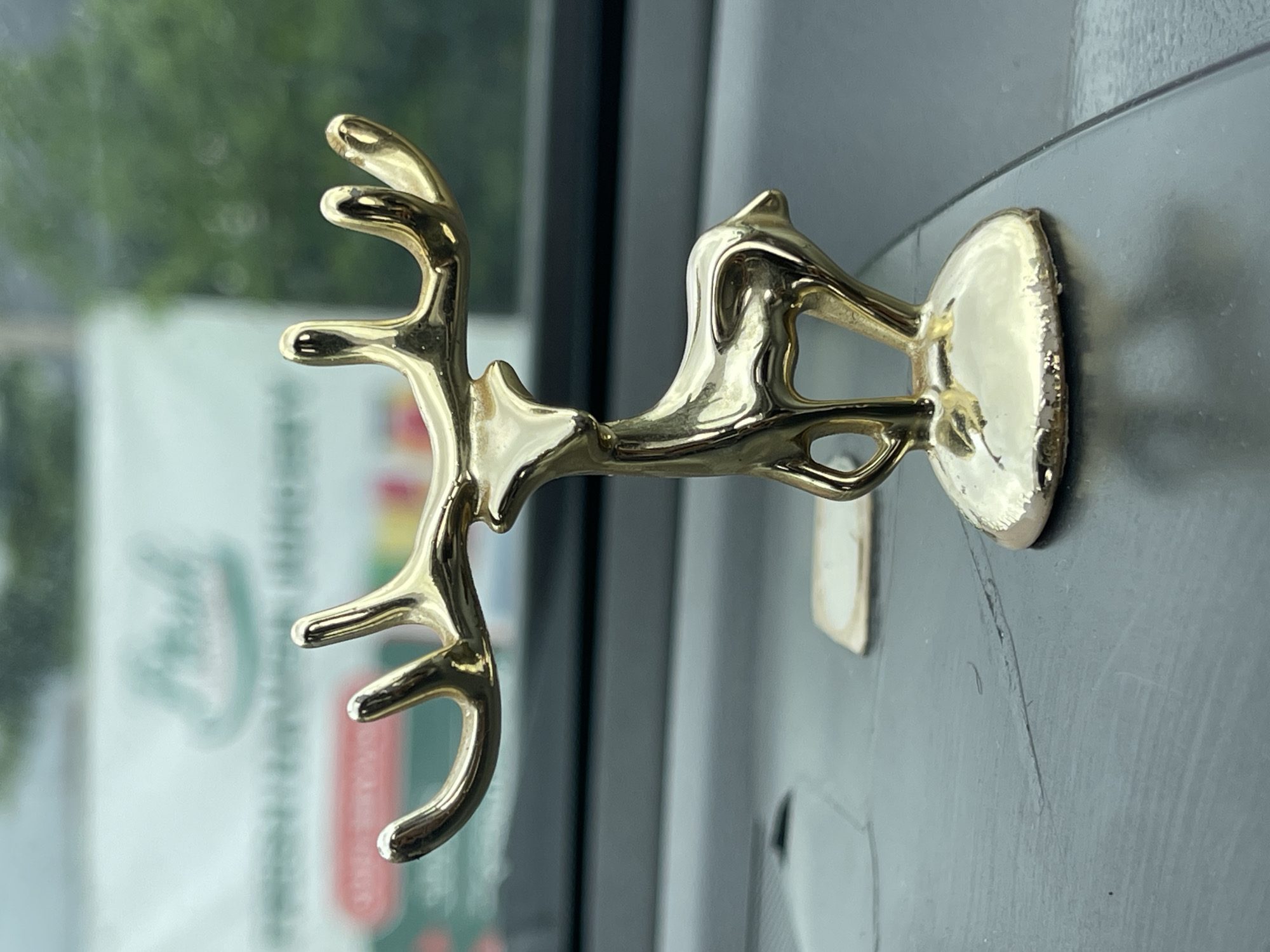 A close-up, eye-level, medium shot captures a small, golden deer figurine standing on a dark, textured surface, possibly a car dashboard. The figurine is positioned centrally, facing left with its head turned slightly towards the viewer. Its antlers are raised and branched, and its body is sleek and stylized, with a polished, reflective surface. The base of the figurine is a rounded, golden disc. In the background, a blurry, light-colored sign with some green and red elements is visible, suggesting an outdoor setting or a view through a window. The overall impression is one of a decorative object placed in a domestic or automotive interior.