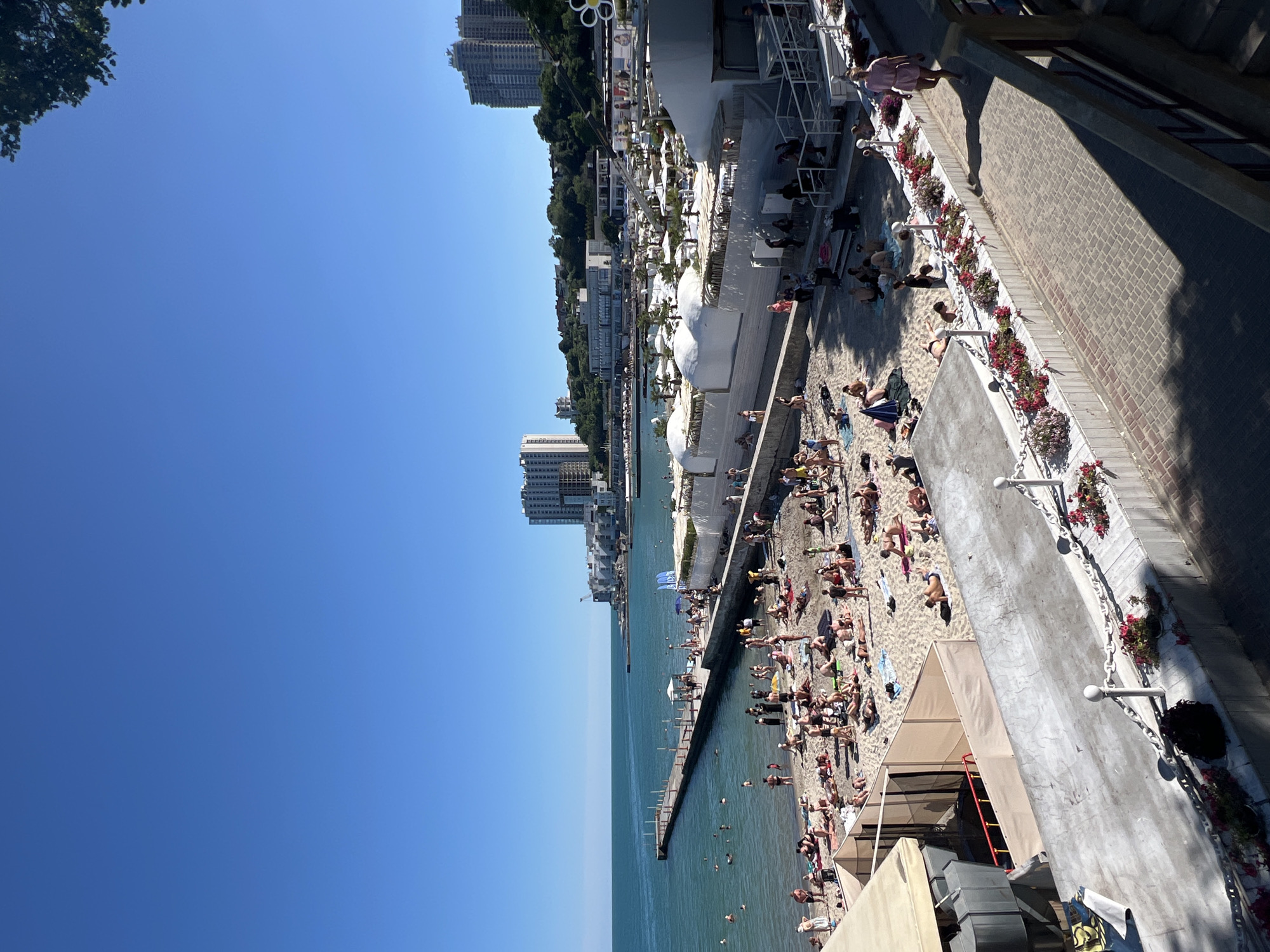 The image depicts a crowded beach scene in Odesa, Ukraine, bathed in bright sunlight, suggesting it is a warm, clear day around midday. People are engaged in typical beach activities: sunbathing on the sand, swimming and wading in the ocean, and relaxing under beach umbrellas and cabanas. The beach is characterized by a stretch of sand leading to a pier that extends into the turquoise water.
Behind the beach, a modern resort area is visible, featuring white domed structures and other beachside amenities. Further inland, taller buildings, some appearing to be apartment complexes or hotels, rise against a backdrop of lush green trees, hinting at the urban environment bordering the coast. A paved walkway or promenade runs alongside the beach, adorned with planters filled with red and pink flowers, providing a decorative element to the seaside setting.
The overall atmosphere appears to be one of leisure and enjoyment, with many individuals casually dressed in swimwear, soaking up the sun and engaging in recreational water activities. The clear blue sky and the bright sunshine contribute to a vibrant and lively scene.
