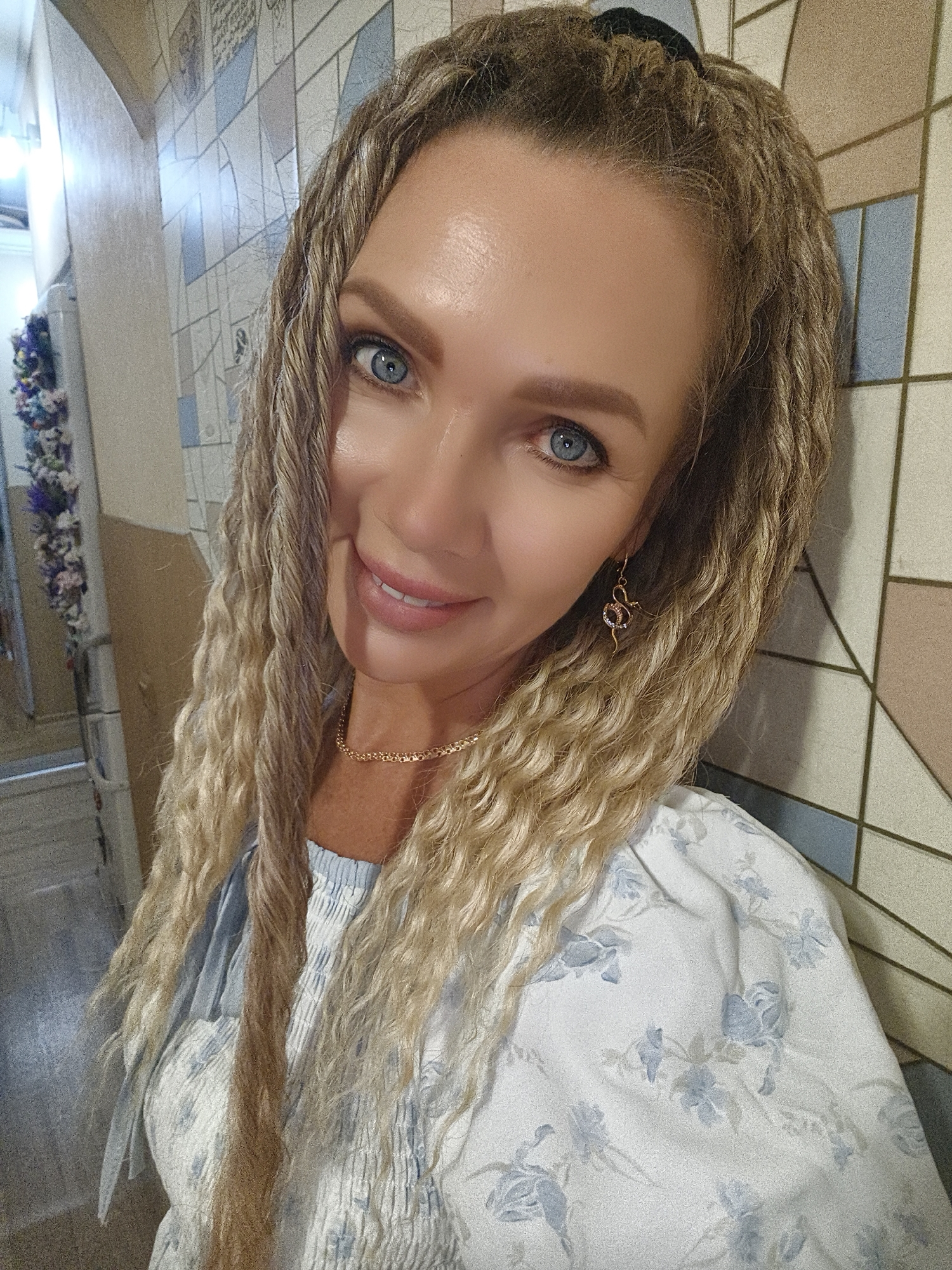 A young woman with blonde braided hair and blue eyes is shown in a close-up portrait. She is wearing a white and blue floral print dress and a gold chain necklace. She is also wearing snake-shaped earrings. She is standing indoors, leaning against a tiled wall. The background shows a hallway with a decorative floral arrangement on the left side. The woman is smiling and looking at the camera.