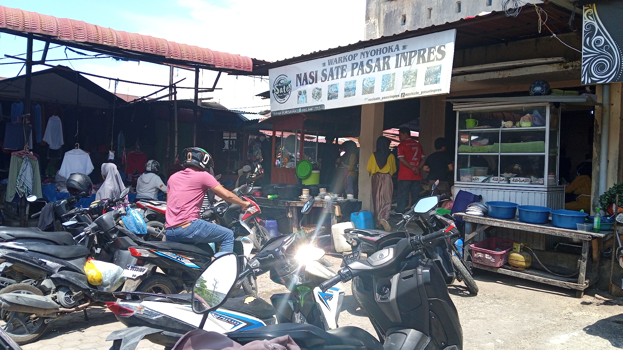 This image captures a street scene in Lhokseumawe, Indonesia, focusing on a small food stall and its surrounding area. The primary subject is a food stall identified by a sign that reads "WARKOP NYOHOKA NASI SATE PASAR INPRES". The stall appears to be a popular spot, with several motorcycles parked in front, indicating customer activity.
In the foreground, several motorcycles are parked, some with riders on them. One rider, wearing a pink shirt and blue jeans, is seated on a motorcycle, looking to the right. Another motorcycle in the foreground has its headlight on, casting a bright light. A person wearing a black helmet is visible on a motorcycle in the middle ground.
The food stall itself is characterized by a glass display cabinet filled with various food items. Behind the counter, several people are engaged in serving or preparing food. A woman in a yellow top and a long skirt is standing, and a man in a red shirt with the number 2 on the back is also present. Another person in black is partially visible behind the counter.
The environment is a bustling outdoor market or roadside area. Stalls with awnings are visible in the background, with clothing items hanging from racks. The ground is paved with concrete. Various objects, including blue plastic buckets, a gas cylinder, and baskets, are arranged on a wooden table in front of the stall. The overall impression is that of a vibrant, everyday scene in a commercial area.