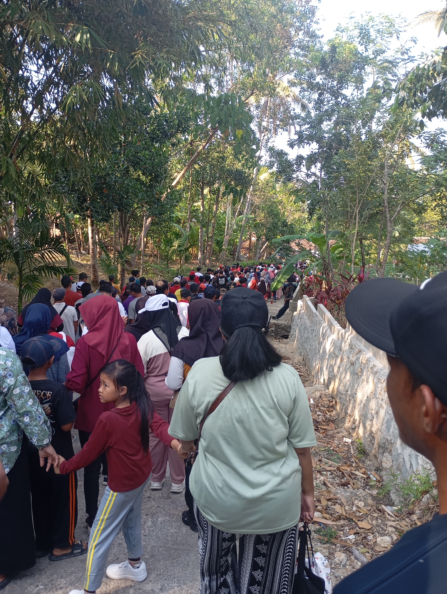 In Ketepung, Indonesia, a diverse crowd of approximately 50-70 people are walking single file along a narrow, unpaved path lined by lush green trees and foliage. The path appears to be a rural or suburban area, with a low stone wall running alongside it. Sunlight filters through the trees, casting shadows on the ground.
The people in the procession are of varying ages and genders, many wearing casual clothing. Several individuals are wearing headscarves or hats. Some are carrying bags. In the foreground, two individuals are partially visible from behind, facing away from the camera and towards the procession. The overall impression is that of a community event or gathering.