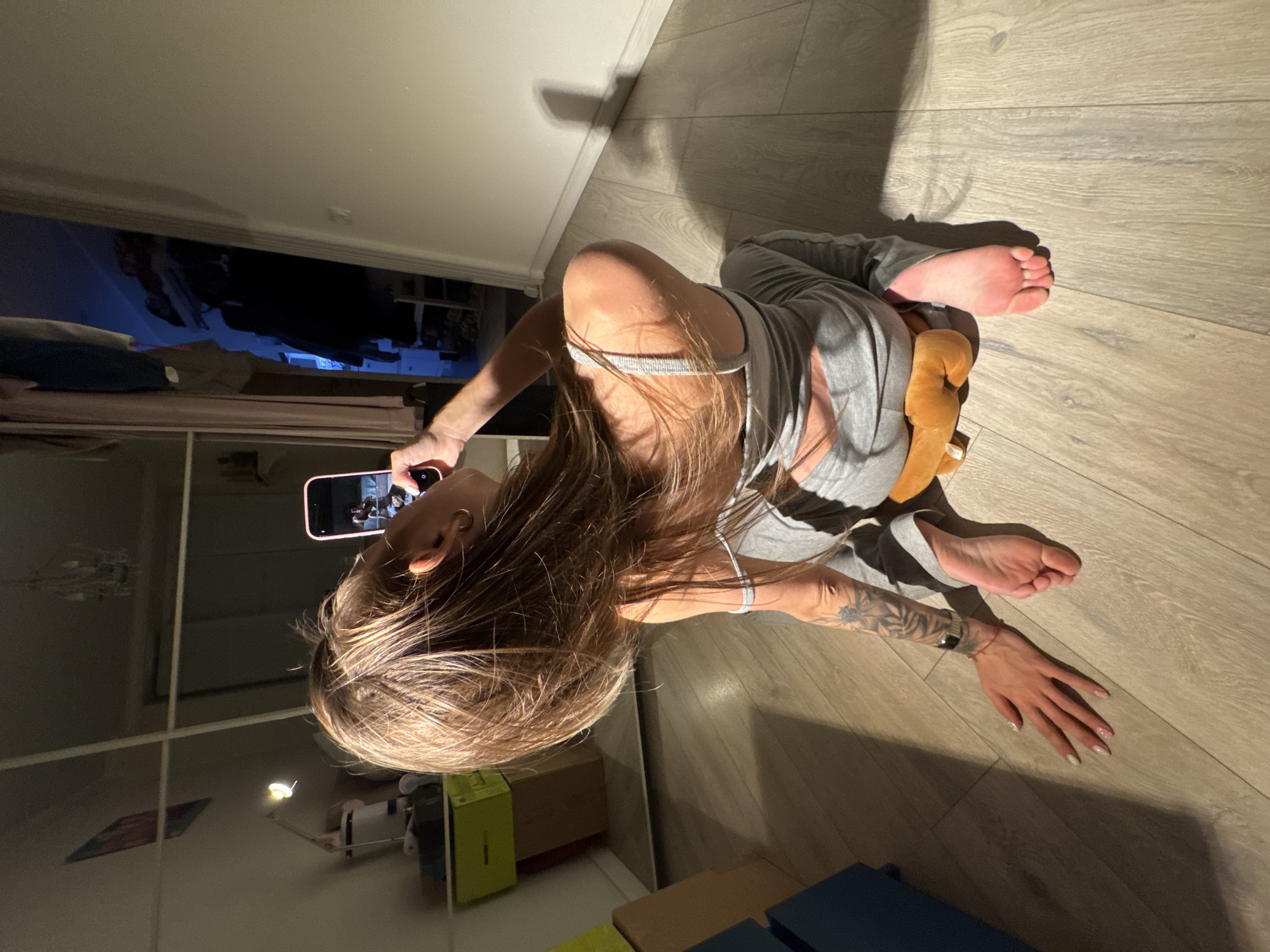 A woman is sitting on a wooden floor, holding a smartphone and taking a photo of herself. She is wearing grey sweatpants and a grey tank top. Her hair is long and blonde, and it falls over her shoulders. She has tattoos on her left arm and a watch on her left wrist. She is barefoot. The room appears to be a bedroom or living room, with a large mirror reflecting a lamp and a chandelier. There are also some clothes hanging in a closet in the background. The lighting in the room is dim, with a strong shadow cast by a window on the floor. The woman is in a contorted pose, with her back arched and her head turned to look at the phone. The scene is forensic and matter-of-fact, with no unnecessary embellishments. The location is the city of Moscow, Russia.