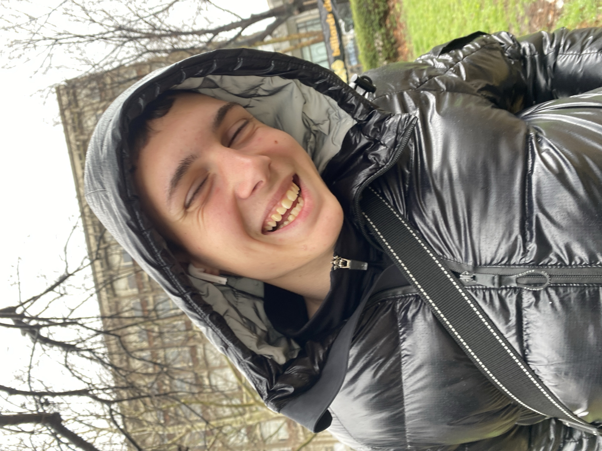 The image depicts a person wearing a black puffer jacket with a hood pulled up. The person is smiling with their eyes closed. The background includes bare trees and a building. Based on the location metadata, the image was taken in Varna, Bulgaria. The specific details of the immediate background are difficult to ascertain.
