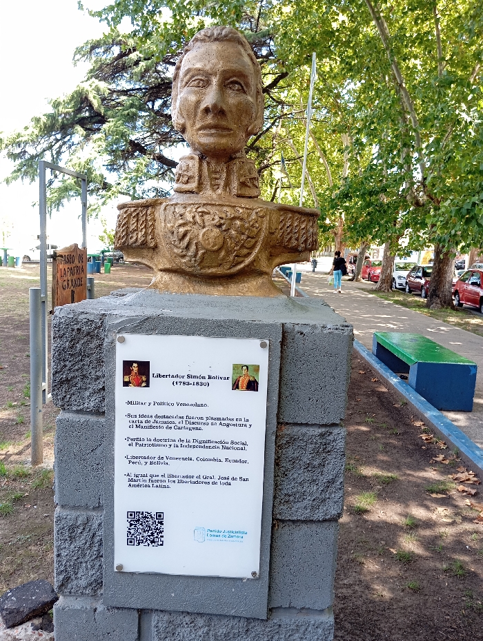 A bronze-colored bust of Simón Bolívar, depicting him in military uniform, is situated on a grey, rough-textured concrete pedestal in a public park in Llavallol, Argentina. A white rectangular plaque is affixed to the pedestal, displaying two small portraits of Bolívar, a QR code, and Spanish text identifying him as "Libertador Simón Bolívar (1783-1830), Militar y Político Venezolano." The text details his key ideas, his role as liberator of Venezuela, Colombia, Ecuador, Peru, and Bolivia, and mentions the "Partido Justicialista Lomas de Zamora" logo. The surrounding ground is a mix of green grass and exposed soil. To the right of the pedestal, a public bench with a blue base and green seat is visible. In the mid-distance to the left, a partial wooden sign reads "PASEO DE LA PATRIA GRANDE." A paved walkway extends into the background, flanked by trees with green foliage. One individual in dark pants and a light shirt walks along the path away from the viewer. A row of parked cars, predominantly red, lines a street parallel to the walkway. The scene is illuminated by natural daylight under a clear sky.