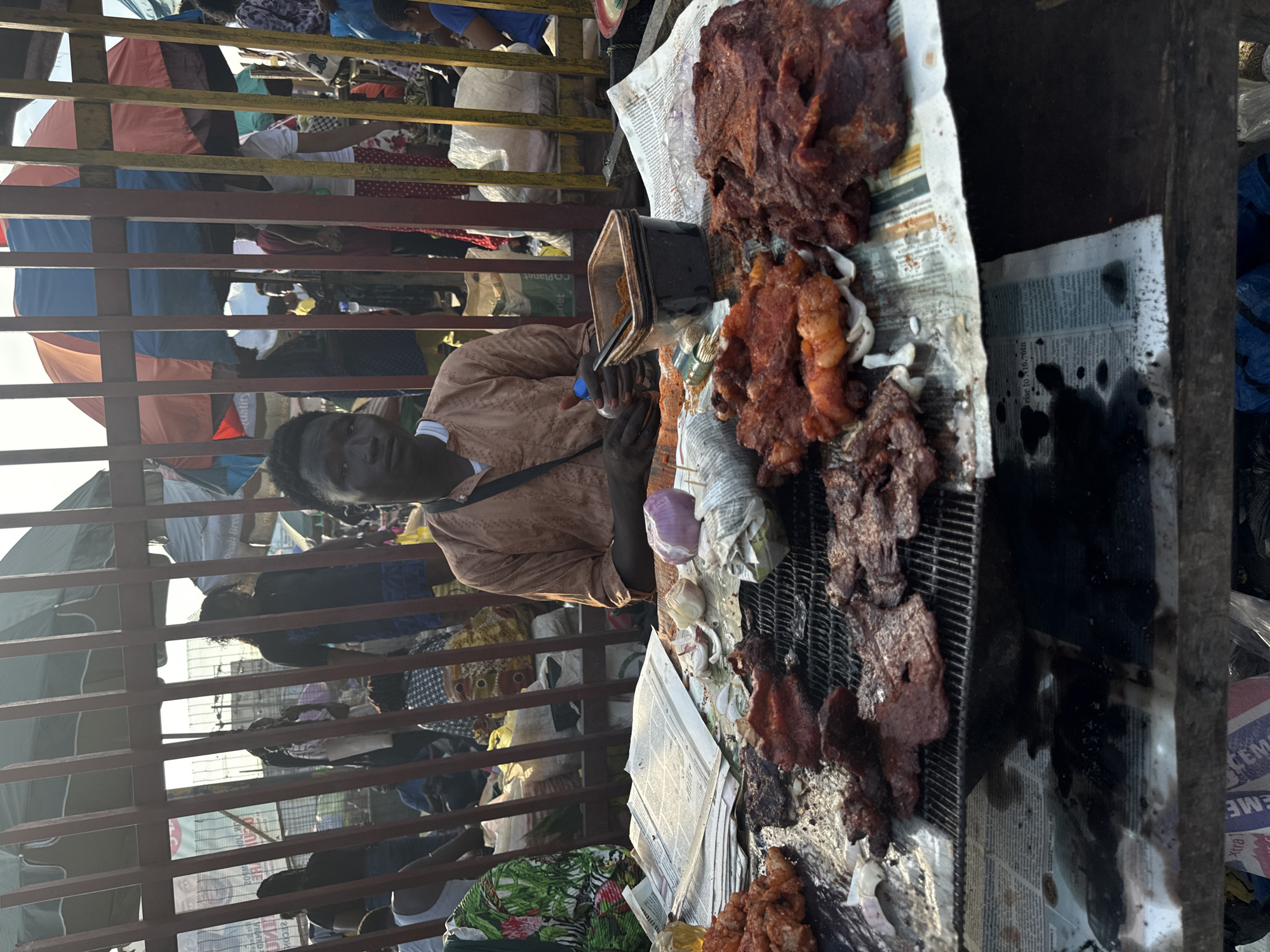 The image depicts an outdoor street food stall within a bustling market environment, consistent with a typical street market in Lagos, Nigeria.
In the foreground, a young Black male vendor, wearing a light brown patterned garment, sits behind a makeshift stall. He looks directly at the camera with a neutral expression. His right hand holds a small blue plastic bottle. The stall's surface is covered with sheets of newspaper, which are stained with oil and residue.
Displayed on the newspaper-covered stall are various cuts of grilled or fried meat, reddish-brown in color, arranged on a dark metal grate. Alongside the meat, a dark brown square plastic container holds a quantity of reddish-brown powdered spice. Scattered on the newspaper are several cloves of garlic, some peeled, and one whole red onion, partially peeled. A small clear bottle with a yellowish liquid and a bundle of small wooden skewers are also visible on the stall.
In the midground, a dark metal fence with vertical bars separates the vendor from a dense crowd of people. Numerous individuals of varying ages and attire are visible beyond the fence, walking and standing within the market. Their faces are partially obscured by the fence and distance.
In the background, numerous large market umbrellas in various colors (red, blue, green, grey, orange) provide shade over other stalls and vendors. Bright daylight illuminates the scene, casting some shadows. Text on a visible newspaper snippet mentions "N16.76tn," indicating the use of Nigerian Naira currency. The overall environment suggests a vibrant, crowded urban marketplace.
