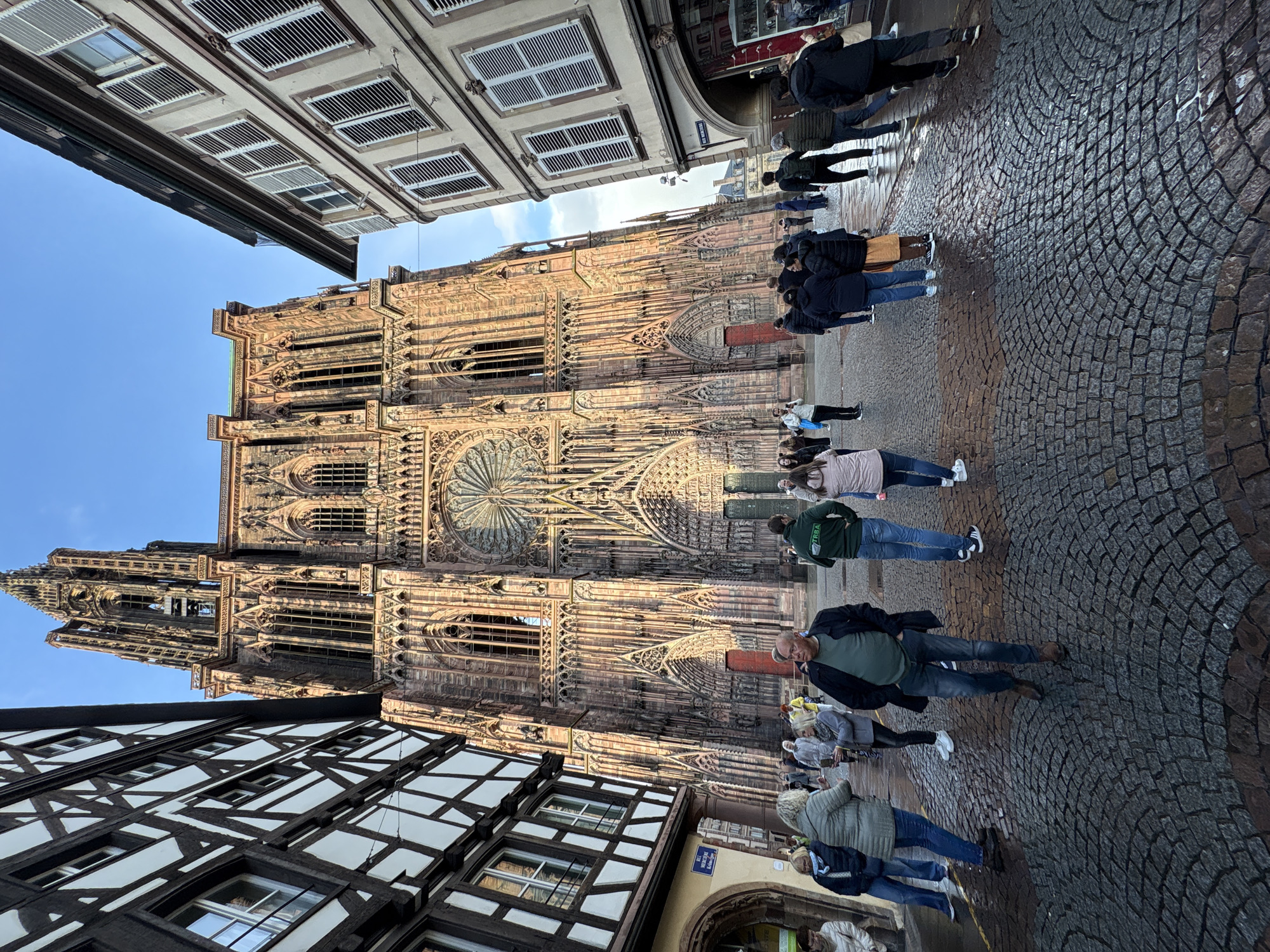 This is a photograph of Strasbourg Cathedral in Strasbourg, France. The cathedral is a large Gothic structure, prominently featuring a rose window. The architecture is ornate and detailed. People are standing on the cobblestone street in front of the cathedral, possibly tourists or locals. To the sides of the cathedral, sections of buildings with typical Alsatian architecture are visible, characterized by half-timbered construction. The buildings have windows and are densely packed with people. The sky is light blue with some clouds. The cobblestone street appears wet, reflecting the buildings and people.