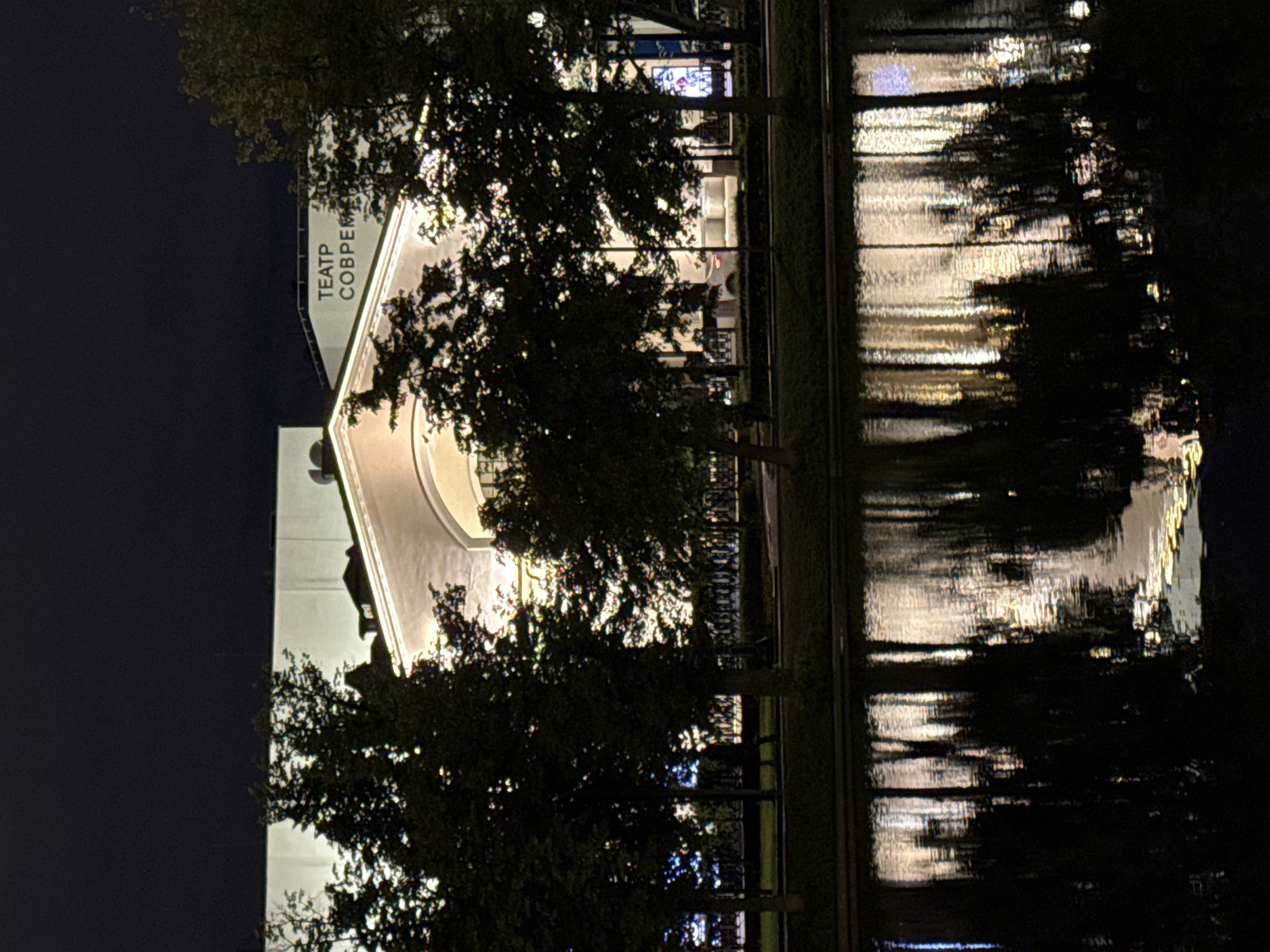 At night, a body of water reflects the illuminated facade of the Moscow theater "Sovremennik," situated in the city of Moscow, Russia. Trees are positioned between the building and the water.