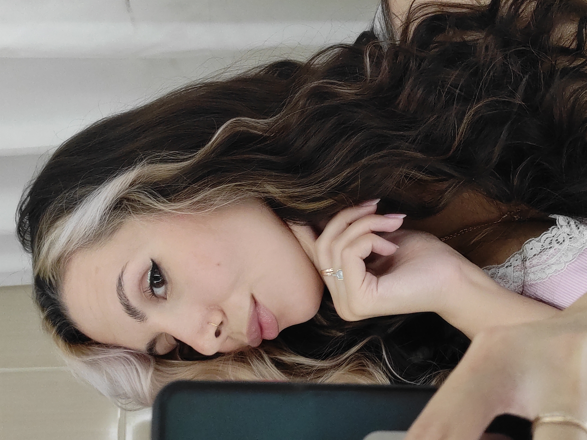 A woman is lying down and appears to be holding a cell phone. The woman has dark hair with blonde streaks near her face, a nose ring, and is wearing a ring. Her shirt has a lace trim. The location is unspecified and there's no information about Chaikovsky, Russia.
