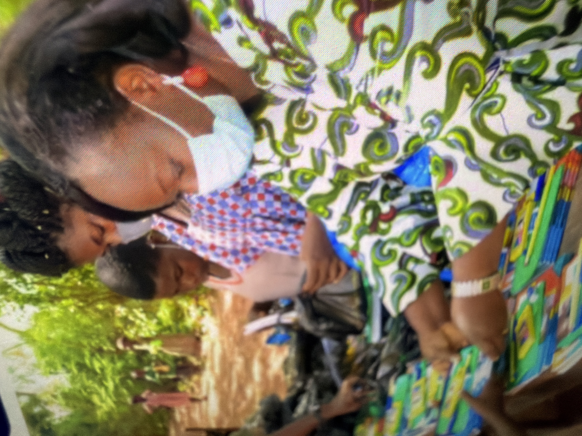 In an outdoor, sunlit environment with lush green foliage in the background, a group of people appear to be engaged in an exchange, possibly distributing or receiving items. Three individuals are prominently featured in the foreground, positioned around a collection of colorful books or pamphlets laid out on a surface.
The person closest to the viewer, on the right, is a woman wearing a light-colored face mask and a vibrant dress with a swirling green and white pattern. She has dark hair styled up and is wearing a prominent orange earring. Her gaze is directed downward.
To her left, a child, also appearing to be wearing a mask, is looking down at the items on the table. This child is wearing a pink and white checkered top.
Further to the left, another person, possibly an adult, is partially visible, with a hand reaching towards the items on the table. This individual is wearing a bracelet with a white band and a green emblem.
In the background, several other figures, including children, are visible in softer focus, suggesting they are further away from the main interaction. The ground appears to be a dirt path. The overall scene suggests an event involving the distribution of educational materials or other supplies in a community setting.