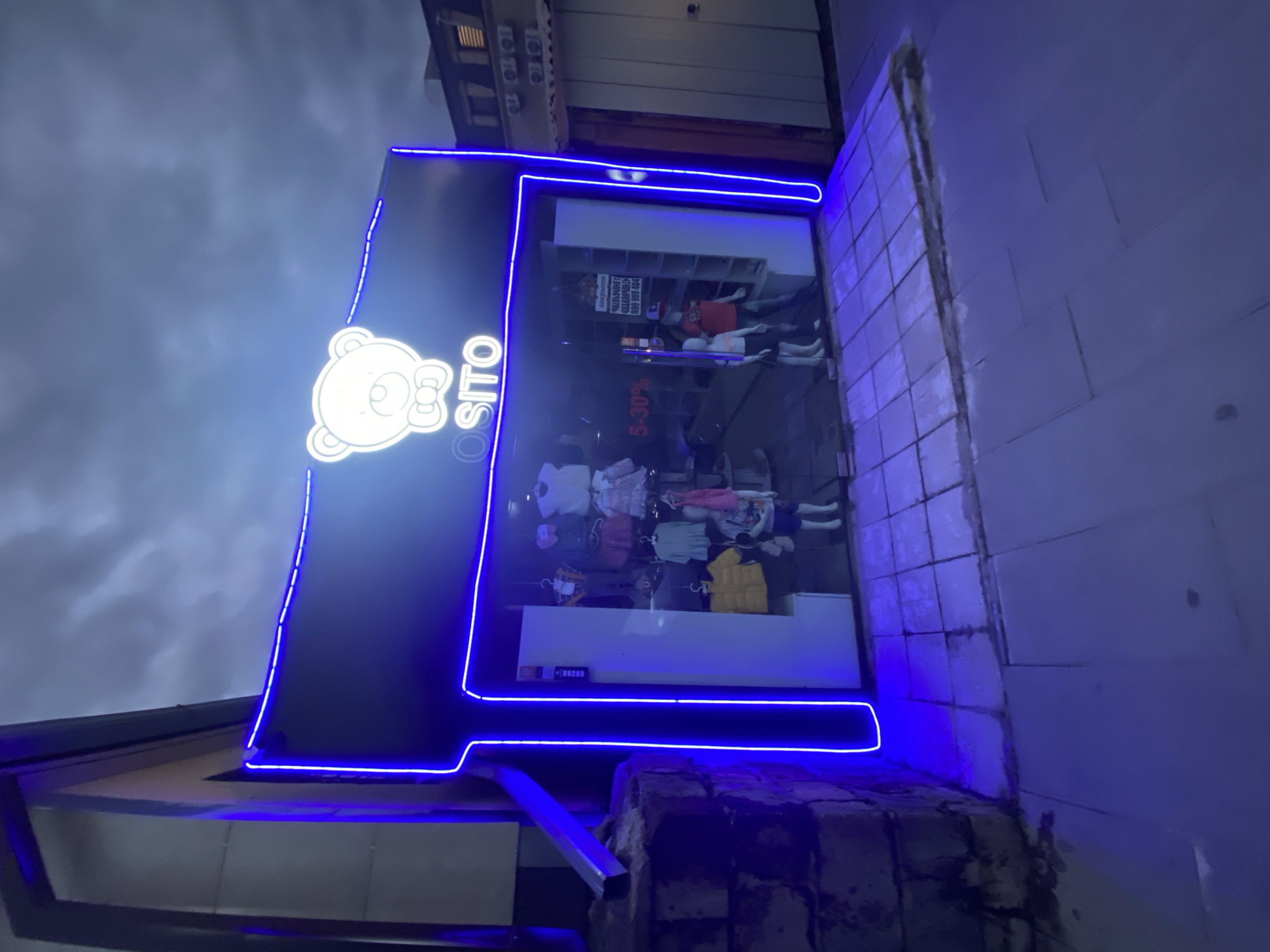 This image captures the brightly lit storefront of a children's clothing store named "OSITO" in Gyumri, Armenia, at an unspecified evening or nighttime hour, under a heavily clouded sky. The storefront is prominently outlined and illuminated by vibrant blue neon lighting, drawing attention to a white neon logo of a teddy bear with a bow tie positioned above the store name.
Inside the glass-fronted store, a display of children's apparel is visible, including dresses, shirts, and jackets on hangers and several child-sized mannequins. Two child mannequins are in the foreground, one in a yellow jacket and the other in a t-shirt and shorts. A larger mannequin in a red shirt and cap is positioned further back inside the store. A "5-30%" discount sign is affixed to the glass, and a white sign inside displays text in a foreign language, including a phone number "099 999 494".
To the immediate left of the store entrance, a metal downspout runs vertically against an adjacent building wall, which transitions into a section of rough, dark-colored stone or brickwork at its base. To the right of the entrance, the building features light brown wooden siding, with an upper section exhibiting multiple external air conditioning units and windows. The ground in front of the store consists of a tiled sidewalk directly abutting the entrance, extending to a wider area of large, lighter-colored square pavers. The blue neon light from the store casts a distinct glow across the sidewalk.