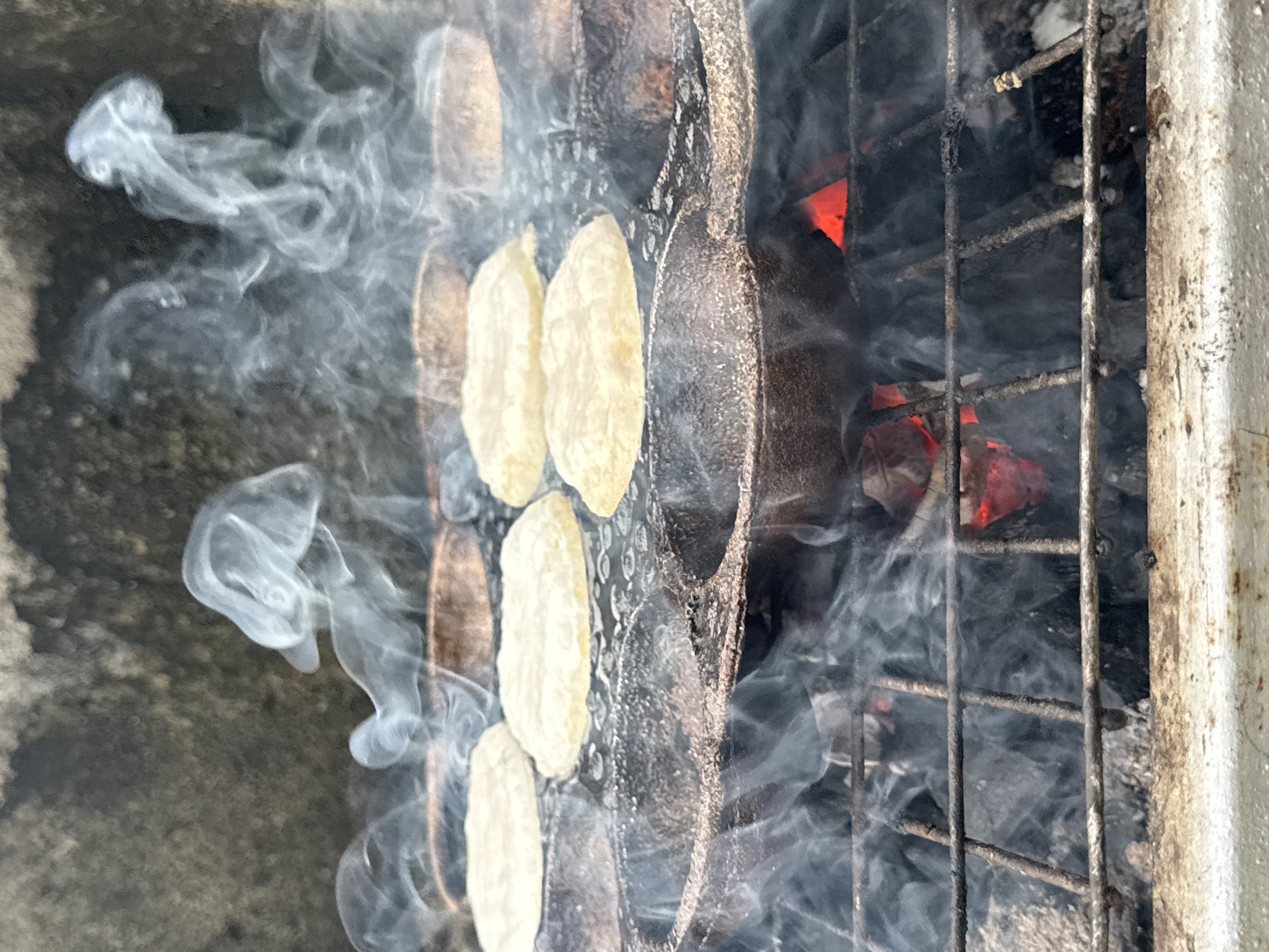 The image shows food being cooked over a charcoal grill. The grill is situated outdoors, possibly in Ado, Nigeria. The food, appearing to be yam or plantain slices, is placed on the grill. Smoke is rising. The grill is made of metal bars. The environment appears to be a concrete outdoor surface. No people are directly visible in the image. The visible actions are cooking and smoking.