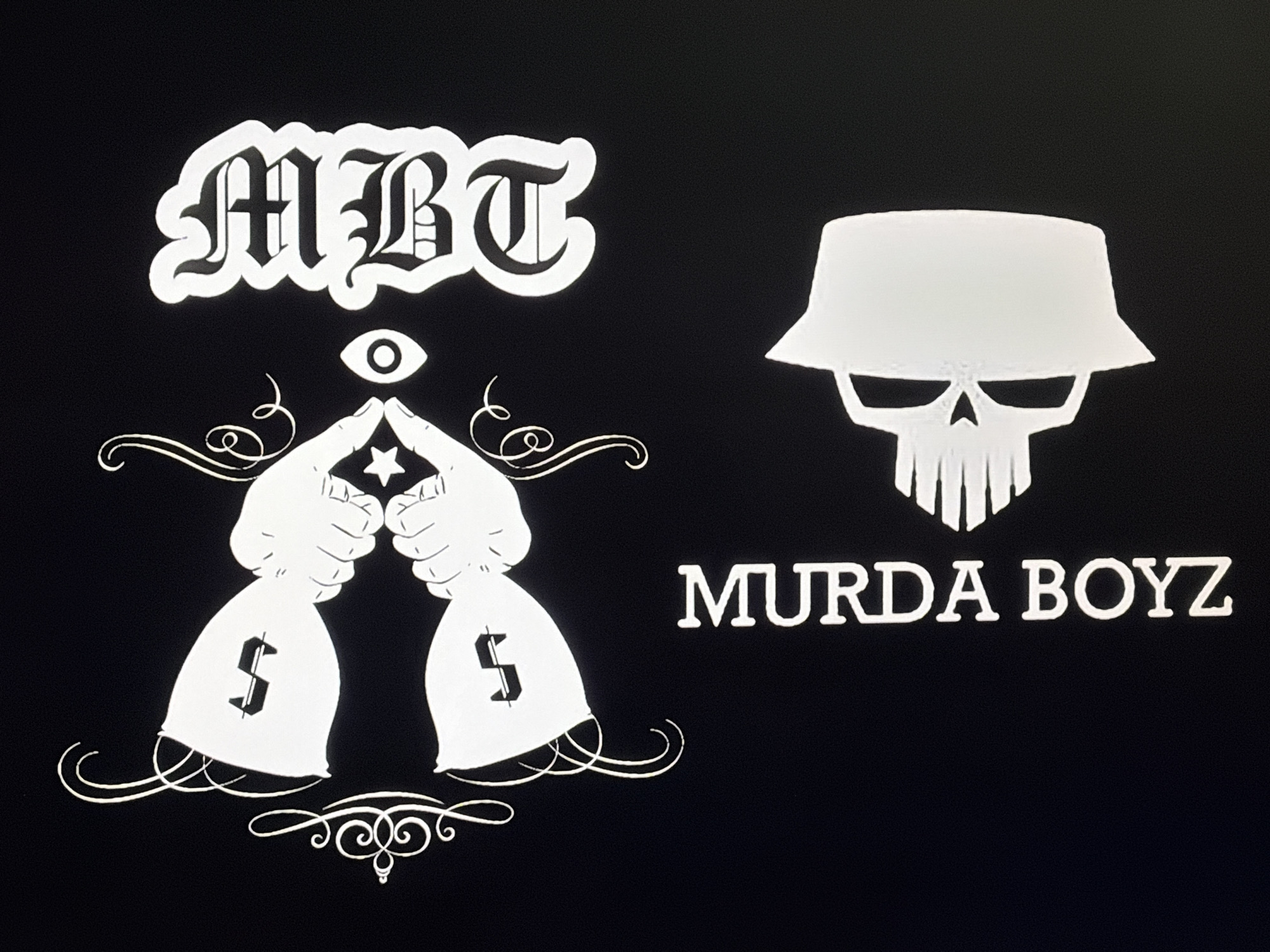 The image is a black background with several white, graphic elements. At the top left, there is lettering that reads "MBT" in an Old English font with a white border. Below "MBT" is an eye, and beneath the eye are two hands, each holding a money bag with a dollar sign. The hands are positioned with index fingers pointing upward toward a star, and decorative flourishes surround the hands and money bags. To the right of these elements is a skull wearing a bucket hat. Below the skull, the words "MURDA BOYZ" are written in a blocky font. There is no information available about a location in Plovdiv, Bulgaria.