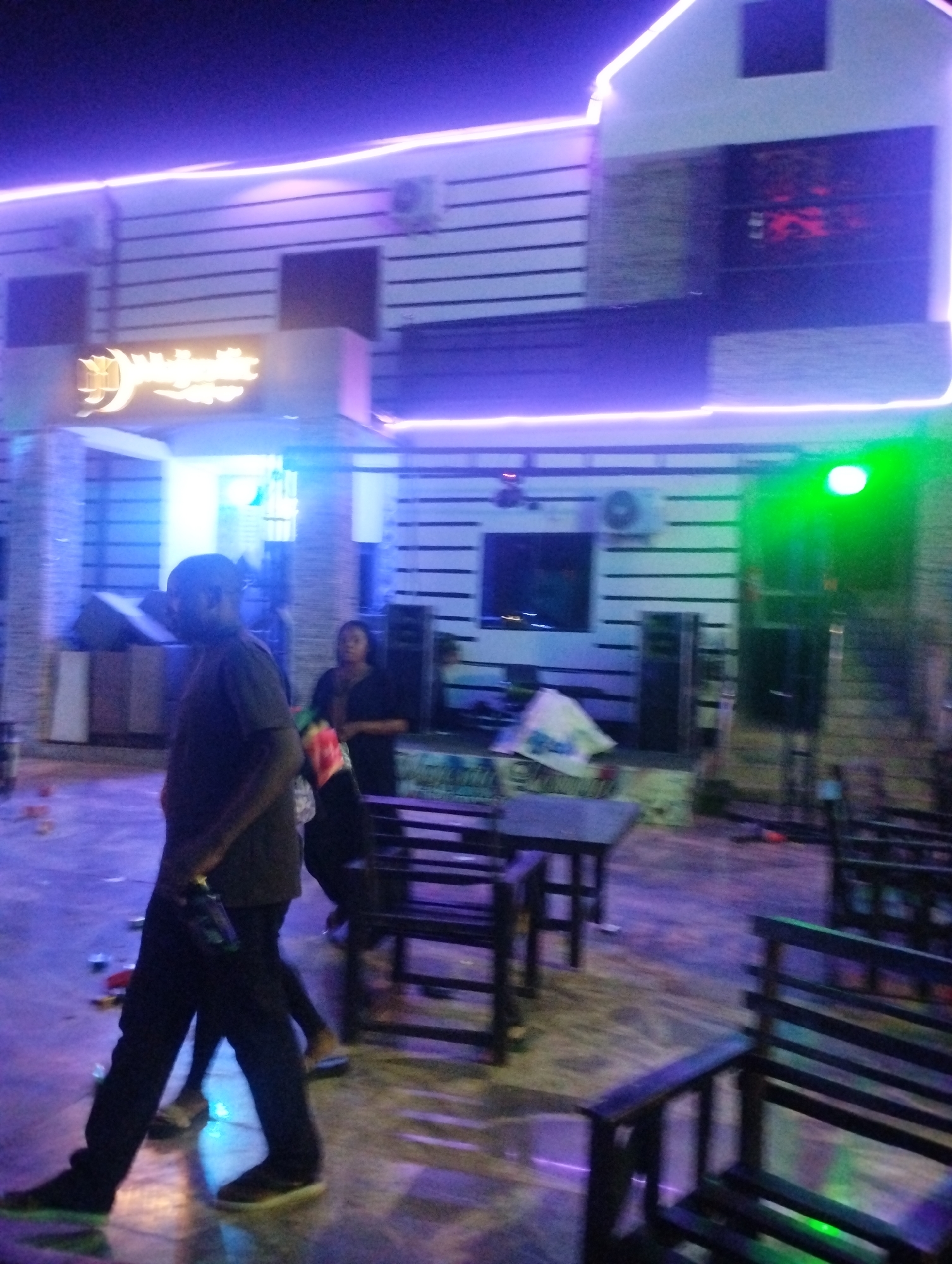 The image shows a building in Tumfure, Nigeria, likely a bar or restaurant, at night. The building has horizontal siding, a sign with illegible writing, and neon lights outlining its shape. A man is walking in the foreground, holding a bottle. Behind him, a woman stands. Several wooden chairs and tables are scattered around the patio area. There are speakers and a television visible against the building wall. The ground is littered with debris.