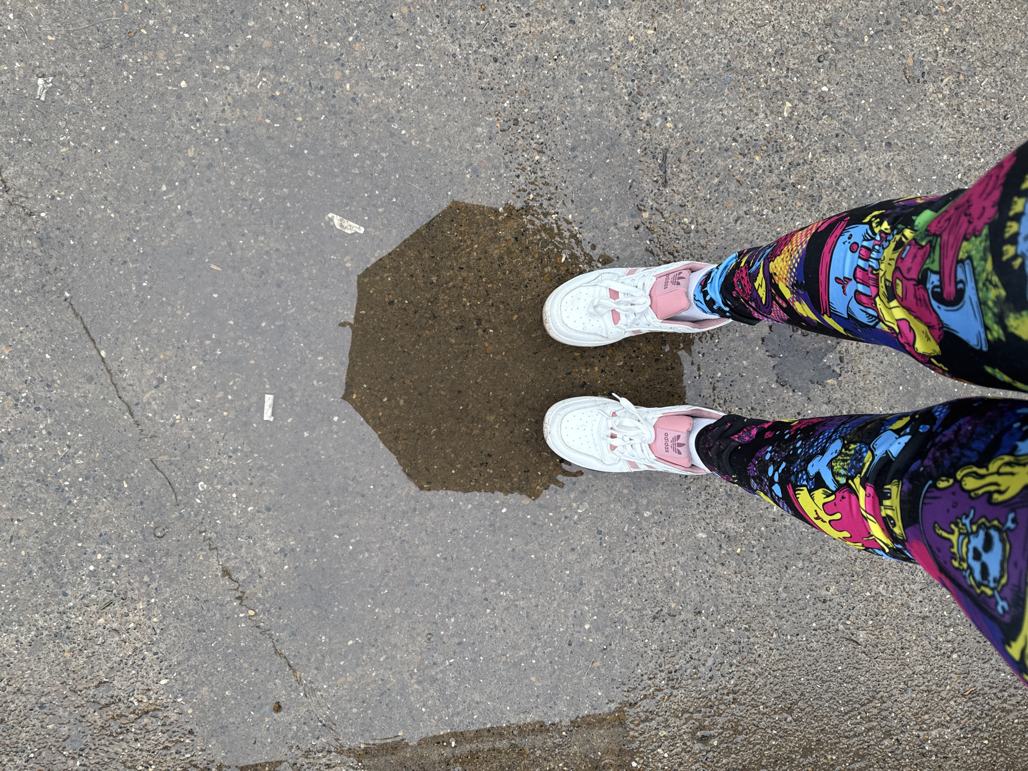 The image shows a person's legs and feet standing on wet pavement. The person is wearing brightly colored leggings with cartoonish designs and white Adidas sneakers with pink accents. There is a puddle of water on the pavement, which reflects an unseen object. The location is not explicitly specified in the image and cannot be reliably ascertained.