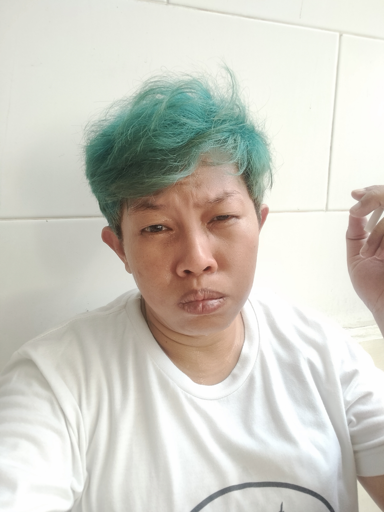 A close-up portrait captures an individual with short, spiky, bright turquoise-colored hair. The person has a medium skin tone and is wearing a plain white t-shirt, which features a faint, dark circular design on the lower left side. Their facial expression conveys a mix of weariness and contemplation, with slightly narrowed eyes and pursed lips. A subtle sheen is visible on their skin, suggesting sweat or oiliness. The individual's right hand is partially visible on the right side of the frame, holding a white, stick-like object, possibly a cigarette or pen. The background consists of a plain, light-colored wall, which appears to be tiled, indicating an indoor setting. The image is captured in Bekasi, Indonesia.