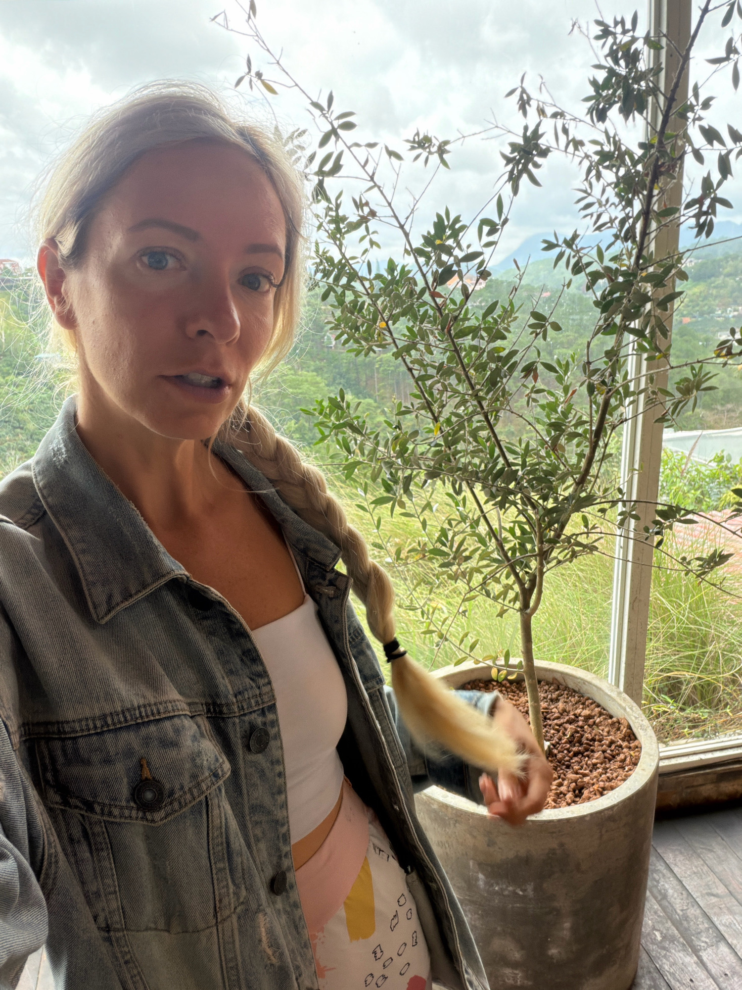 A young Caucasian woman with blonde hair in a braid, wearing a white tank top and a denim jacket, stands indoors near a large potted plant. She is looking at the camera with her mouth slightly open. Behind her, through a window, a lush, green landscape with hills and mountains can be seen, consistent with the terrain of Da Lat, Vietnam. The cloudy sky suggests overcast weather. The woman appears to be in a residential setting or a cafe with large windows overlooking the natural scenery.