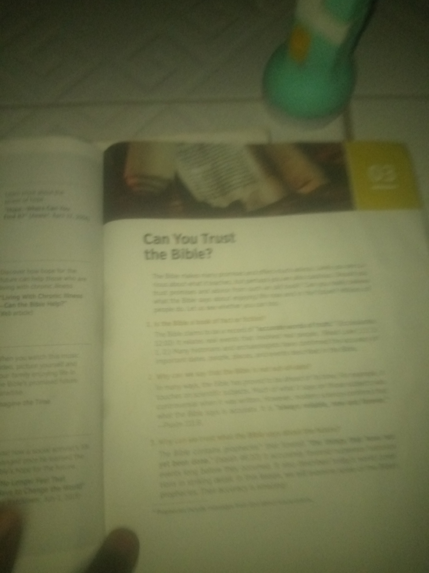 The image shows an open book with text and an illustration on the pages. The text on the right page asks, "Can You Trust the Bible?". The left page contains a list of article titles. The environment appears to be indoors, with a blurred object, possibly a lamp, in the upper right corner. The image is blurry and overexposed, making it difficult to discern fine details. There is no information in the image that can be used to determine its location in Jauro Yinu, Nigeria.
