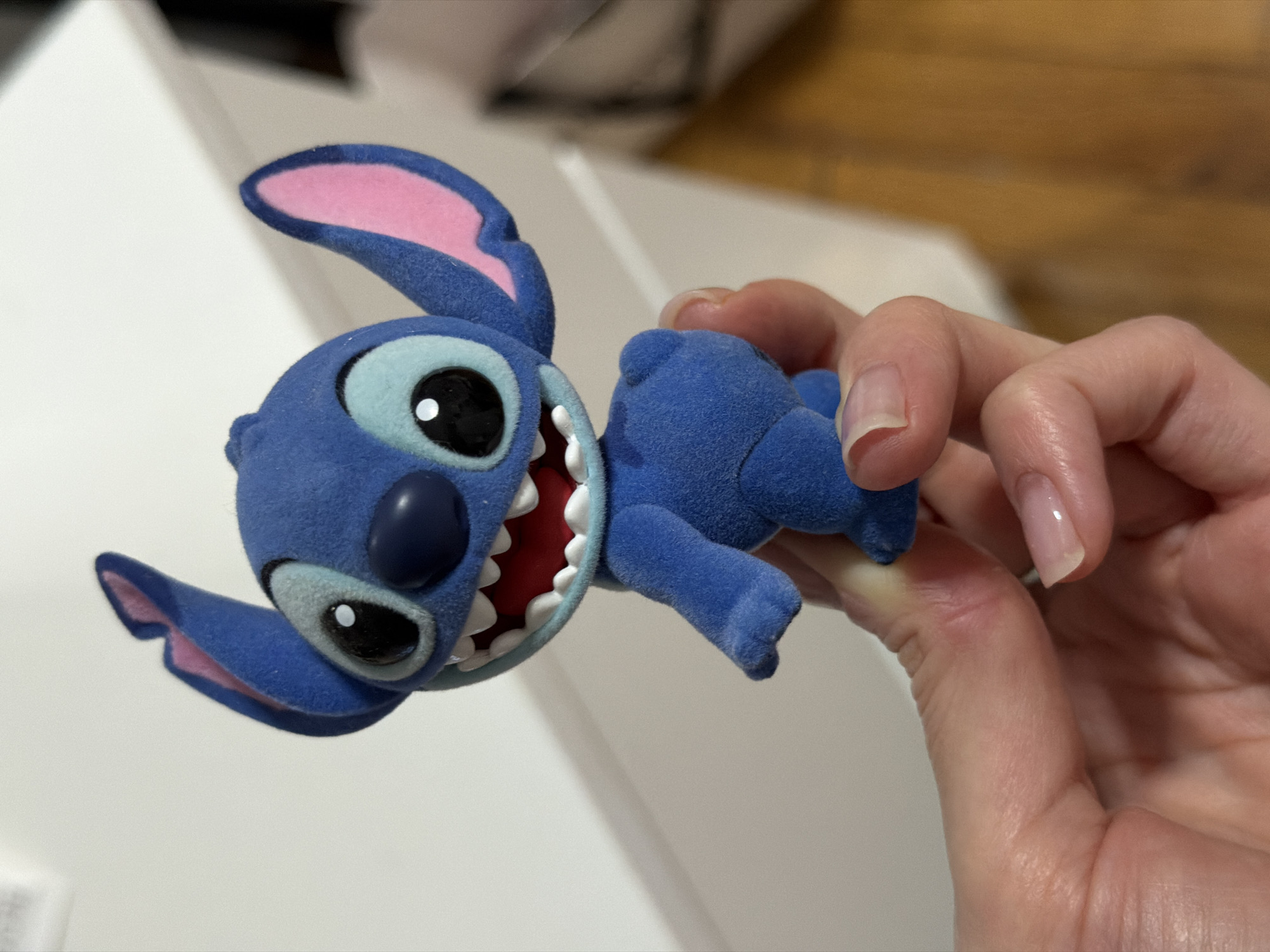 A blue, fuzzy figurine of the Disney character Stitch is held in a person's hand. The figurine has large, round eyes with white pupils, large pointed ears with pink insides, and a wide mouth with sharp white teeth and a red interior. Stitch's body is blue, with a small tail and two small arms. The hand holding the figurine has light-toned skin with visible fingernails. The background is blurred, showing a white surface and a wooden surface. No location in Moscow, Russia is identifiable.