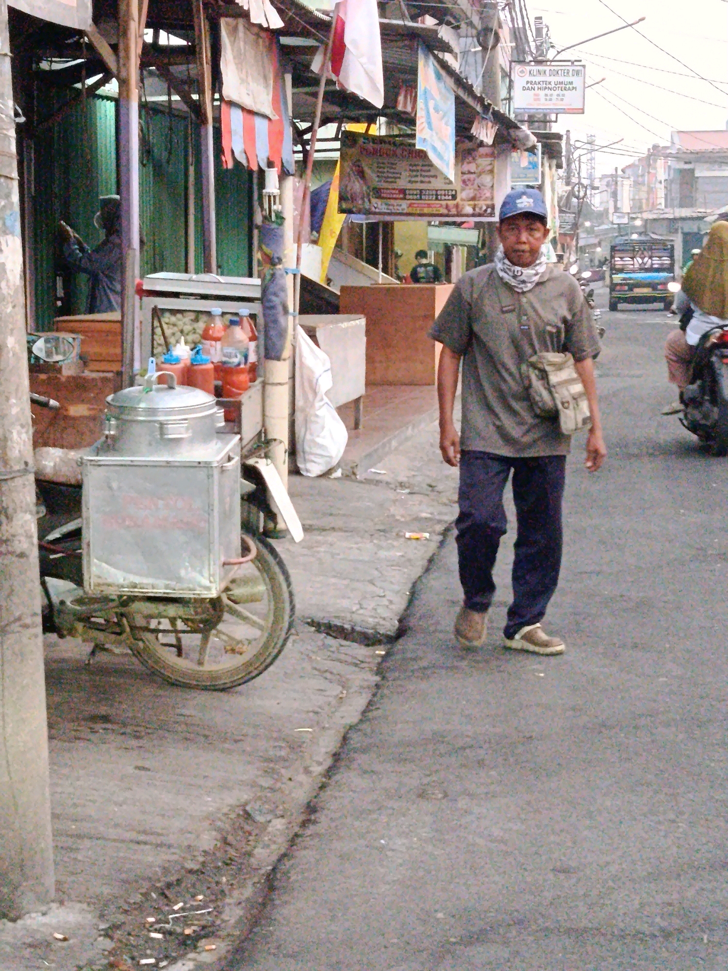 A man walks on the right side of a street in Bekasi, Indonesia, while a street vendor's cart is parked on the left. The man wears a blue cap, a gray t-shirt, dark blue pants, and brown shoes. He carries a tan messenger bag over his shoulder. The street vendor's cart, attached to a motorcycle, has a large stainless steel container on top. Behind the cart, bottles of sauce and other condiments are visible. Several shops and buildings line the street, with awnings and signs indicating commercial activity. In the background, traffic can be seen, including a small truck. The overall scene depicts a typical street view in a commercial area of Bekasi.