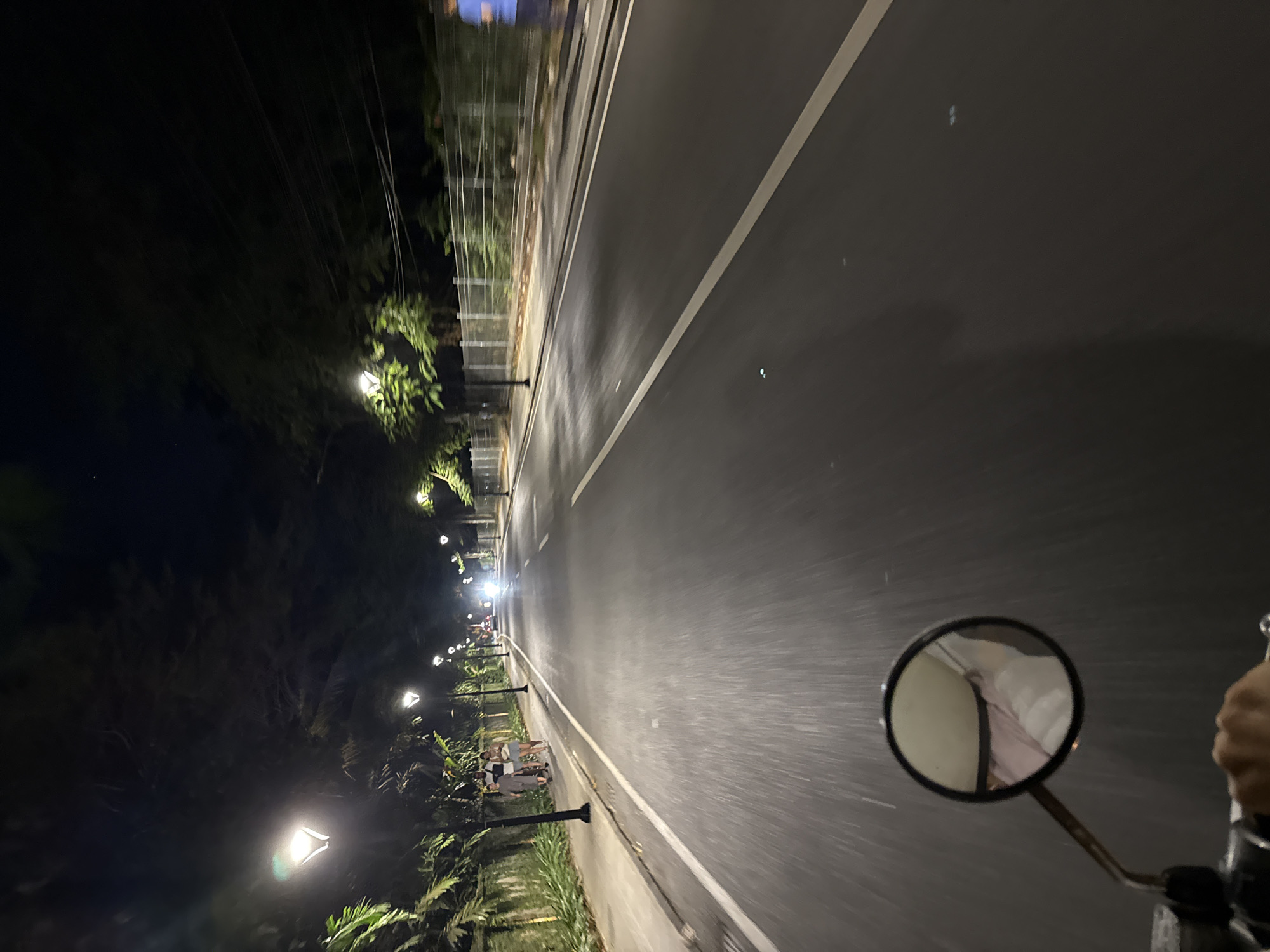 The image depicts a nighttime scene on a paved road, likely in Denpasar, Indonesia, given the visual cues. The perspective is from a moving vehicle, possibly a motorcycle, as indicated by the visible rearview mirror in the foreground. The road is illuminated by streetlights, with several visible along the left side of the street, casting a warm glow. The road itself has distinct white lane markings. On the left side of the road, there is a sidewalk lined with lush green vegetation and a low wall. A few pedestrians are visible on the sidewalk. On the right side of the road, there is a fence and more vegetation. The overall atmosphere is quiet and serene, typical of a well-lit urban street at night.