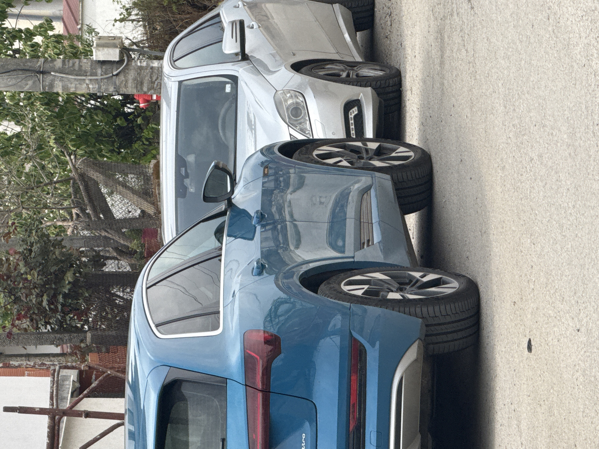 A blue Audi and a silver Mercedes are parked on a street in Parvomai, Bulgaria. The Audi is in the foreground, angled towards the viewer, displaying its rear quarter panel, tire, and taillight. Behind it, the silver Mercedes is partially visible, with its front quarter panel, tire, and headlight noticeable. The vehicles are parked on an asphalt surface. A grey power pole is positioned to the left of the silver car. In the background, a chainlink fence, trees, and parts of buildings are observed.