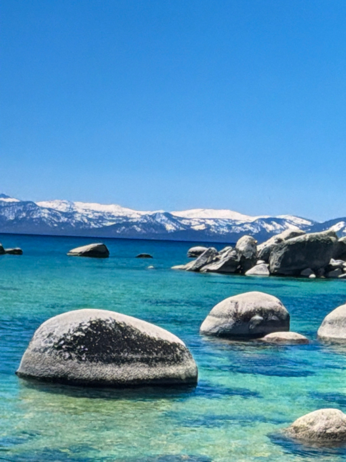 This is a landscape photograph featuring a body of water with large rocks. The water is clear and turquoise, with visible ripples and reflections. Several large, smooth, grey rocks are partially submerged in the water. In the background, snow-capped mountains are visible under a clear blue sky. The scene evokes a sense of natural beauty and tranquility. The location is not Kafr Yasif, Israel.